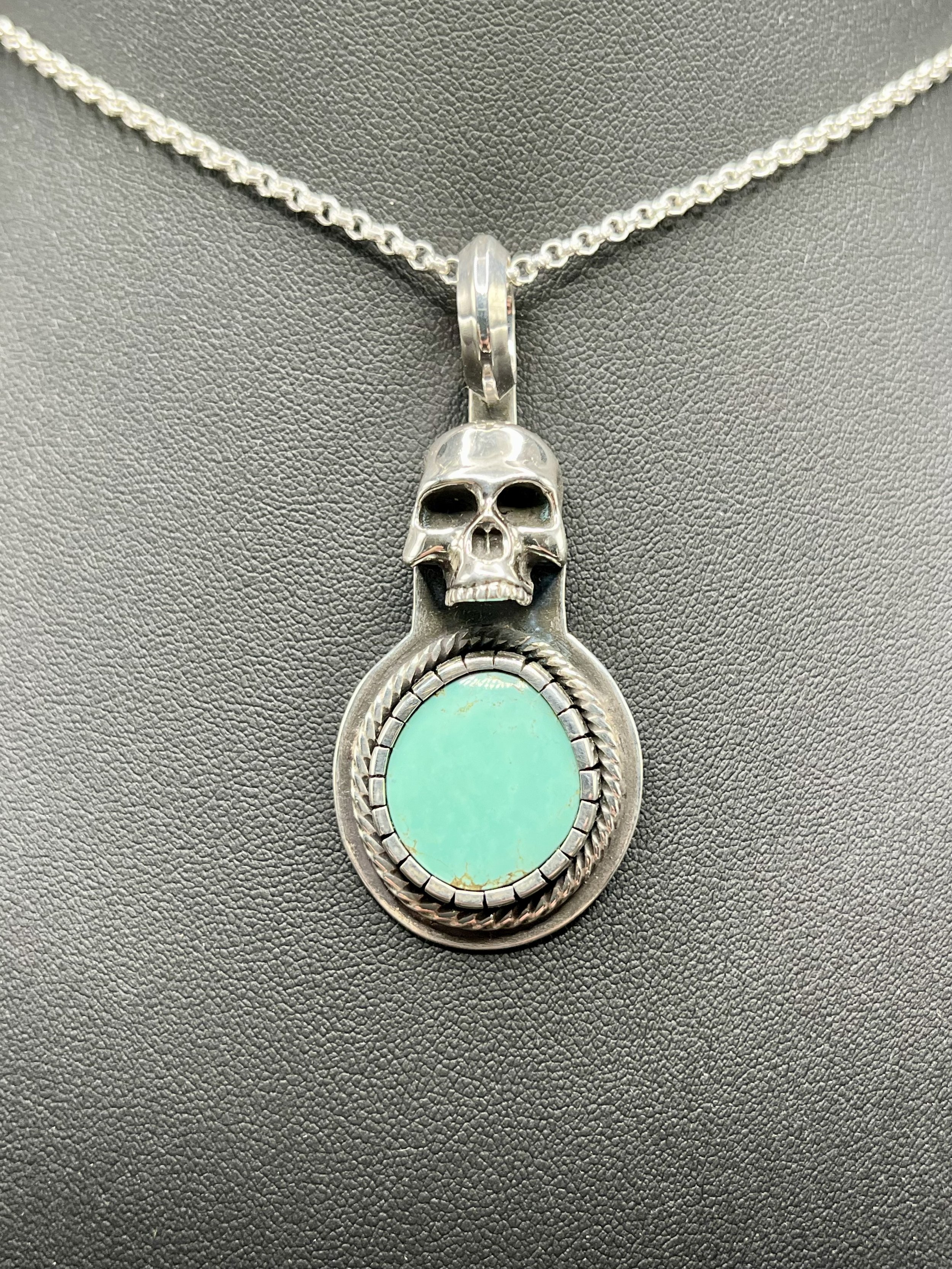 A silver necklace with a skull pendant and bright turquoise gemstone displayed on a black mannequin.