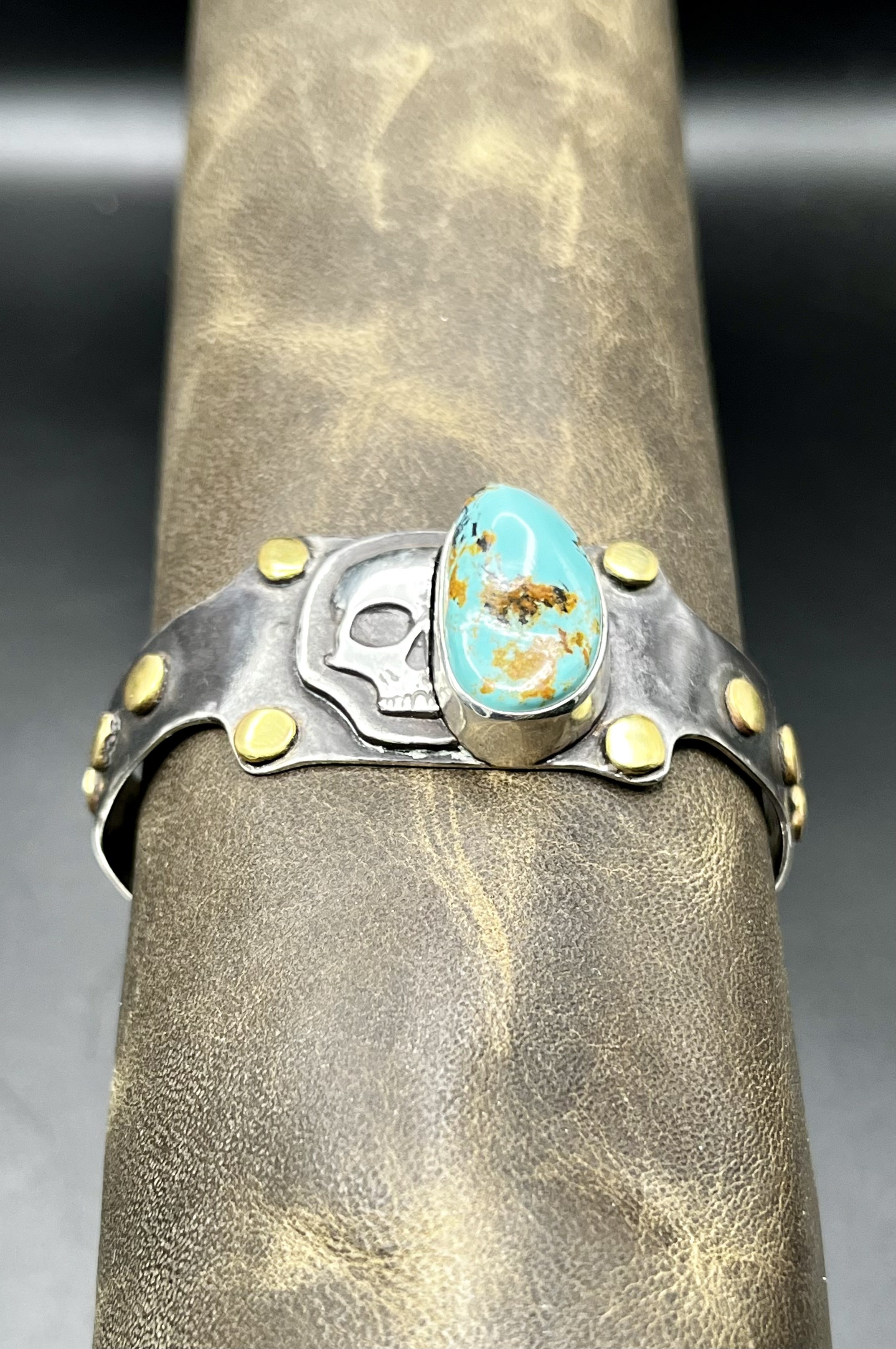 A sterling silver bracelet with gold beads and a skull in the middle, with a large Kingman turquoise nugget covering half of the skull's face. The bracelet is wrapped around a mannequin's arm.