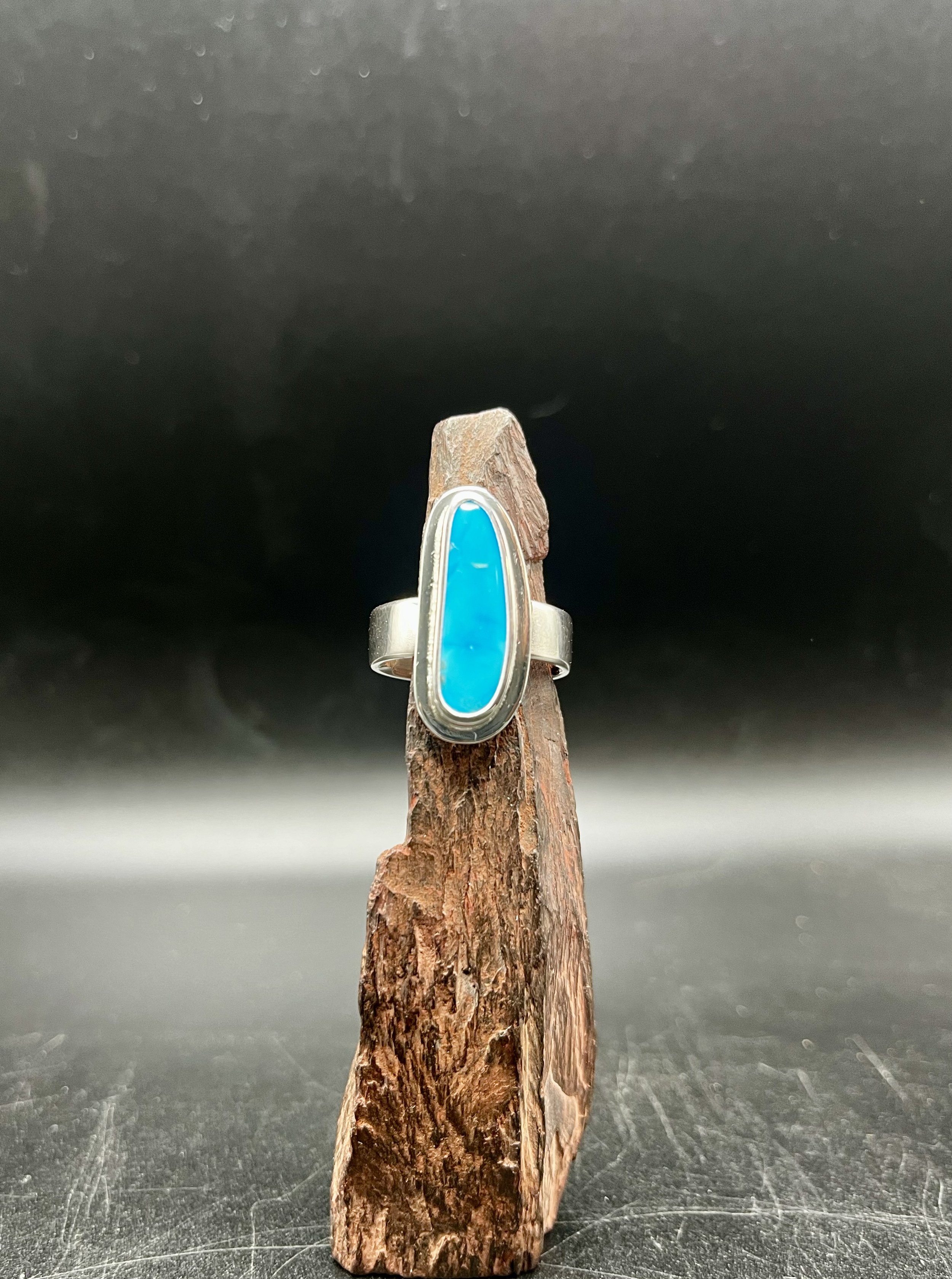 The Sleeping Beauty Turquoise Sterling Silver Ring, custom-designed by KC Silversmith, displayed on a jagged, rust-colored rock in a black room. The rock is rested on a black display stand.