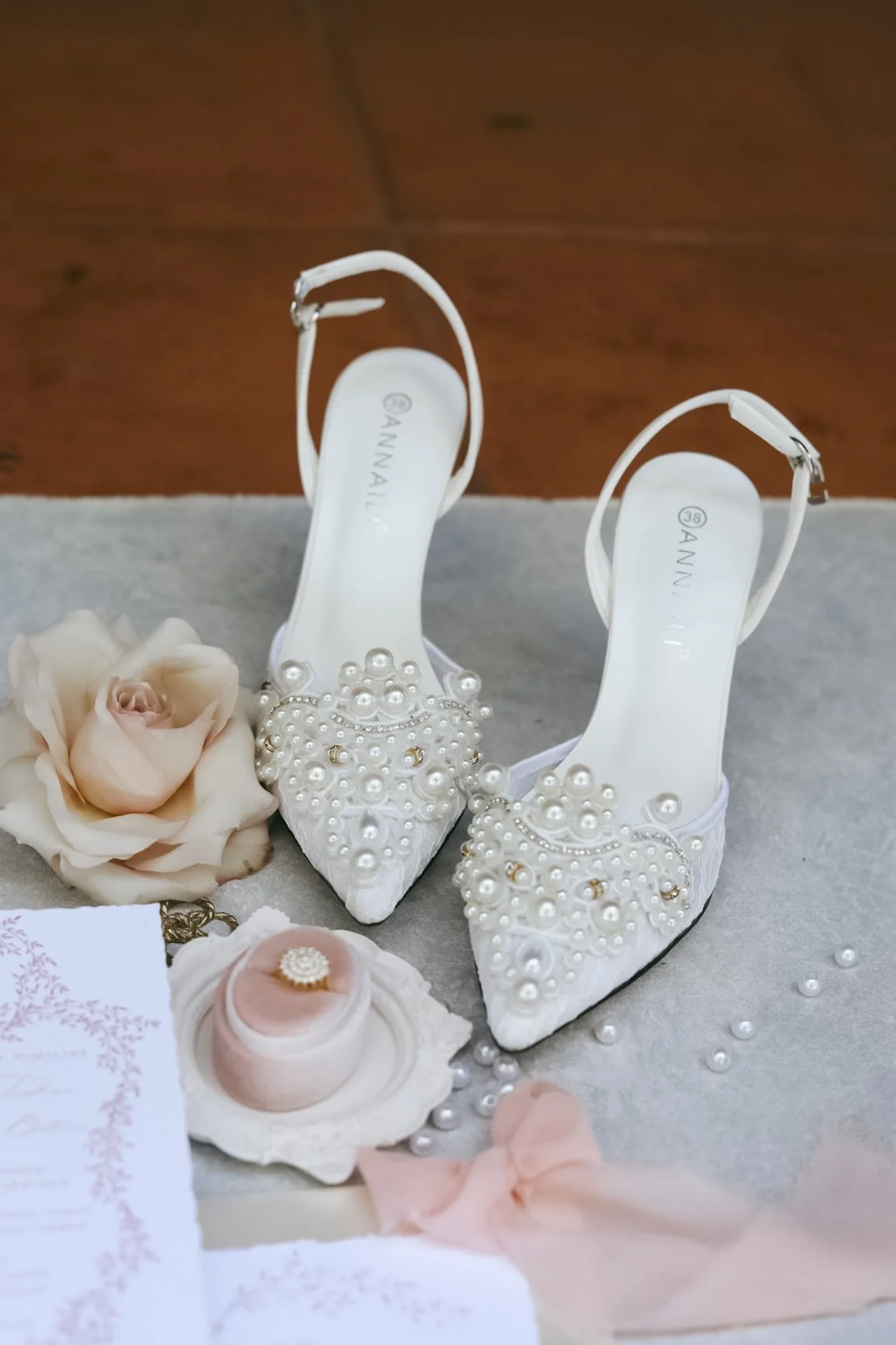 Bridal accessories with embellished high heels, a pink ribbon, a flower, a wedding ring in a box, and wedding invitations.