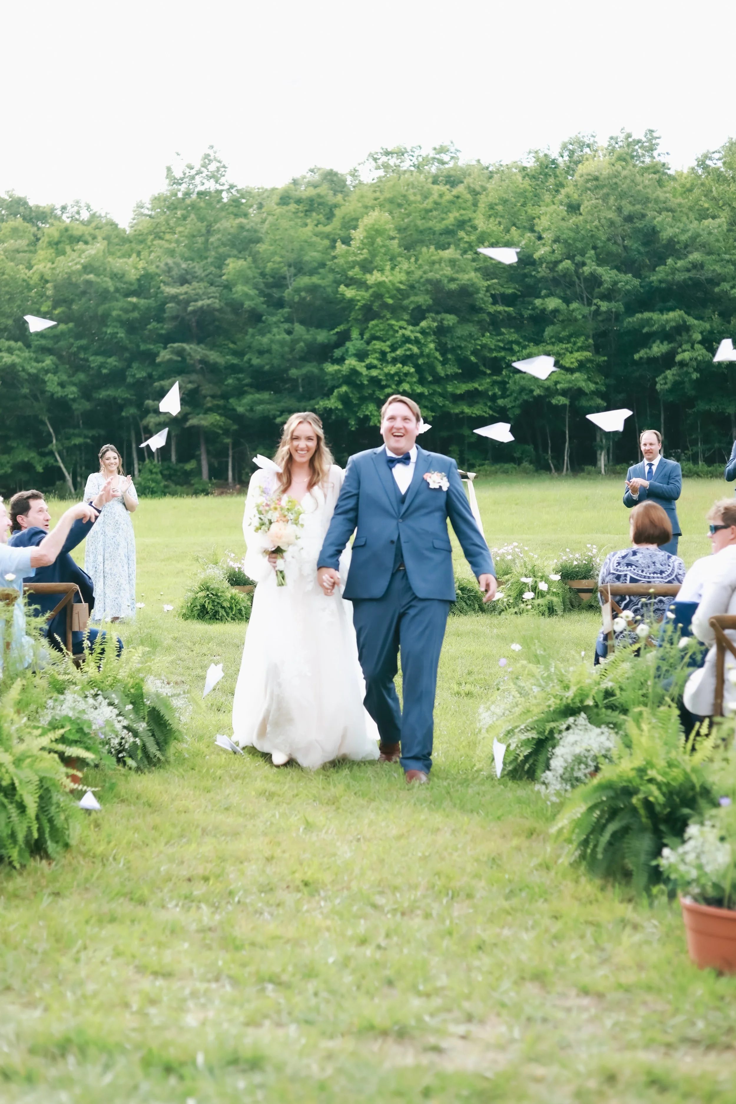A bride and groom walking down a grassy aisle during an outdoor wedding ceremony. They are smiling and holding hands, with the groom in a blue suit and the bride in a white gown holding a bouquet. Guests are seated on either side, throwing paper airp