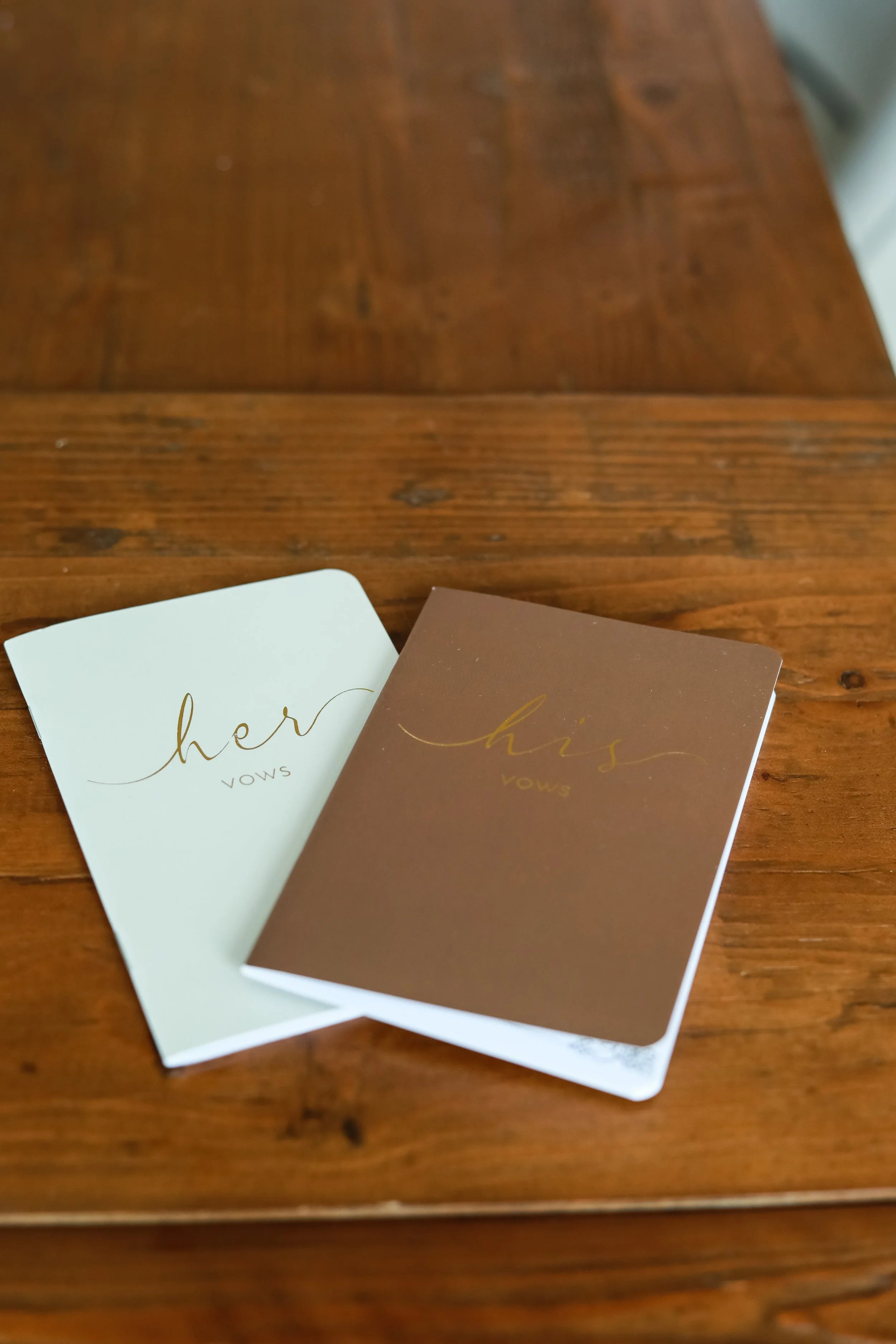 A pair of vow books labeled "her vows" and "his vows" on a wooden table.