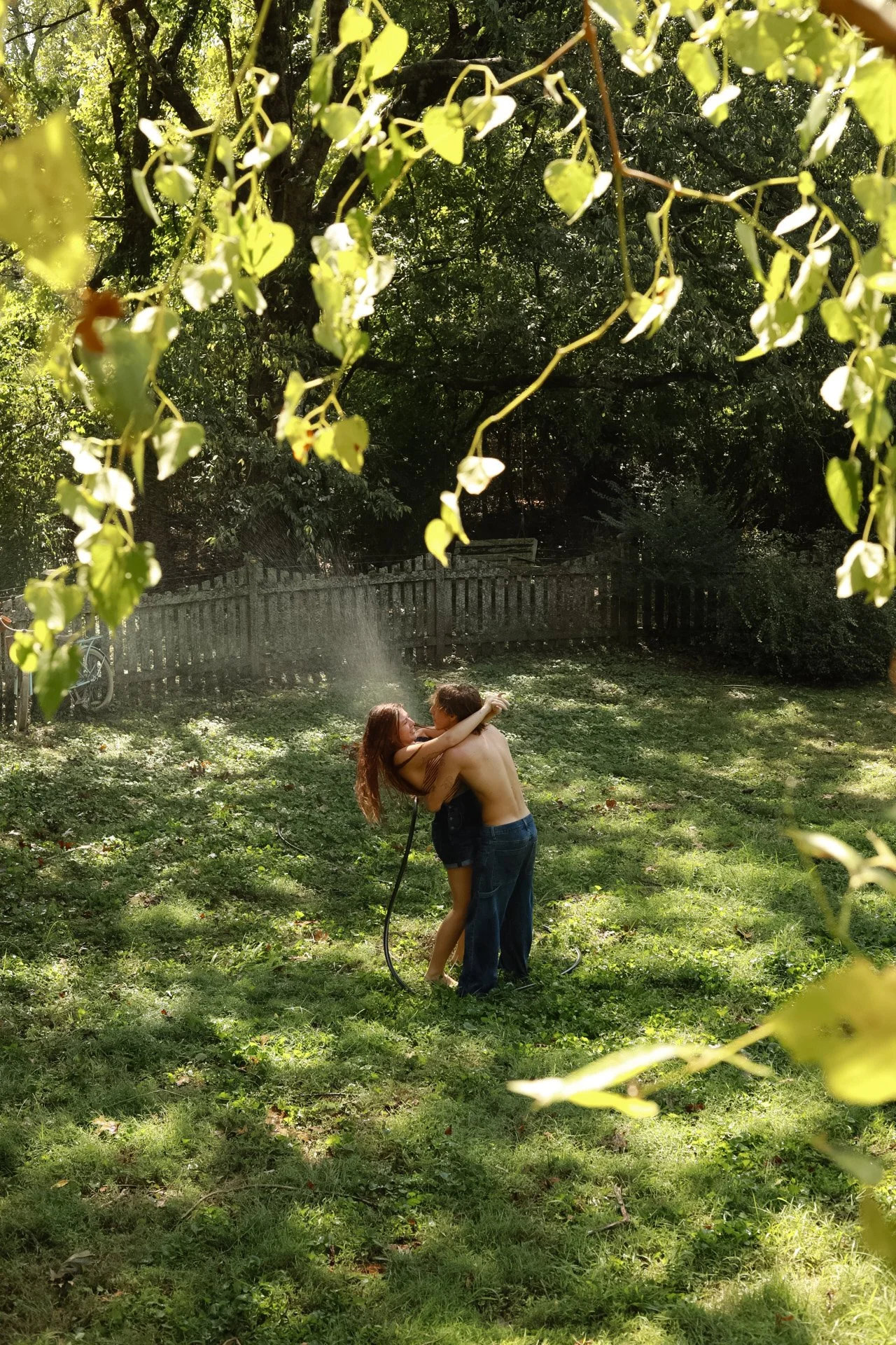 A young couple dancing and embracing in a backyard garden, surrounded by green grass, trees, and a wooden fence. The scene is shaded with sunlight filtering through the leaves.