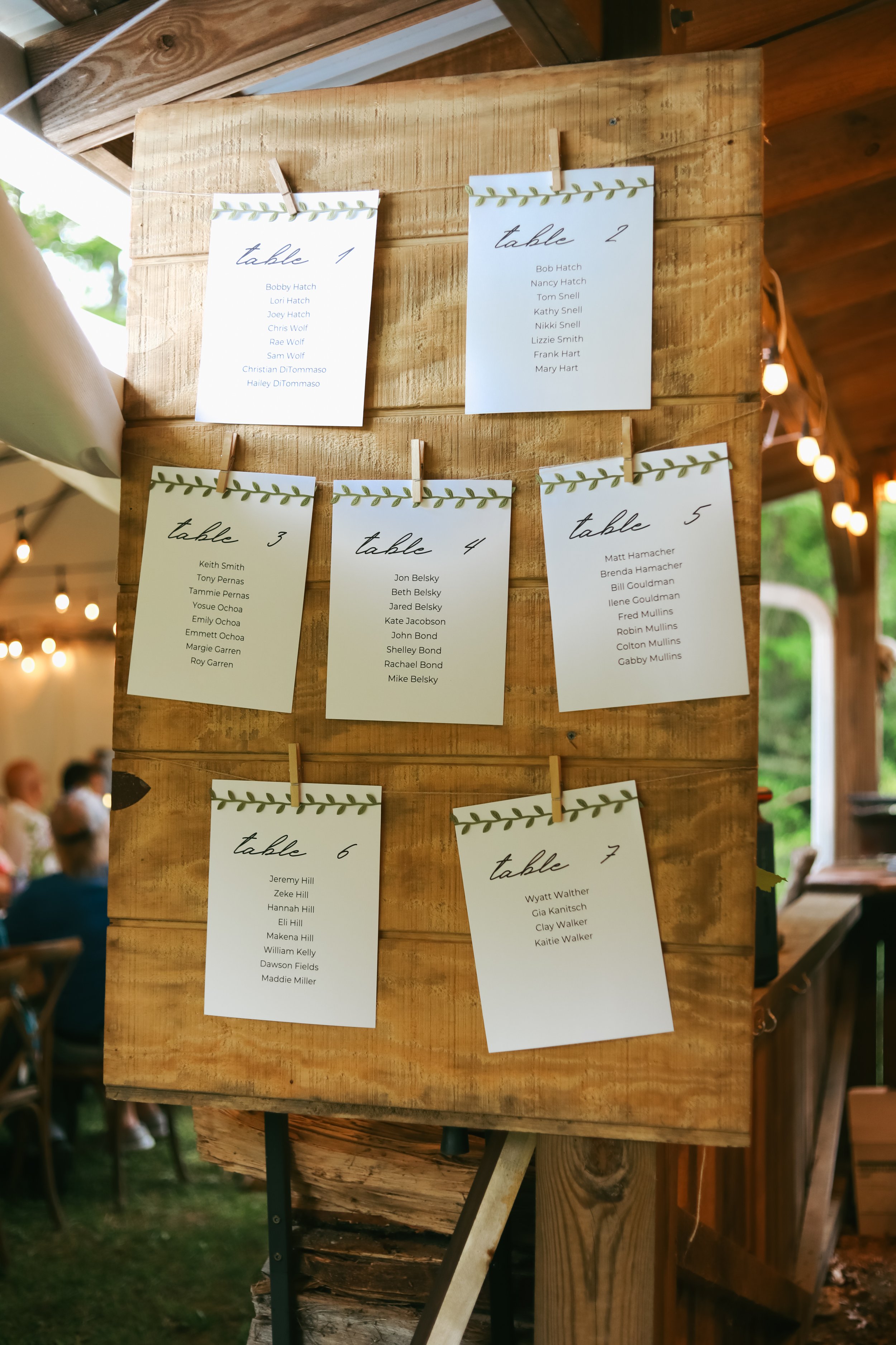 Wedding seating chart with table assignments displayed on a wooden board, featuring individual table lists with names clipped to the board.