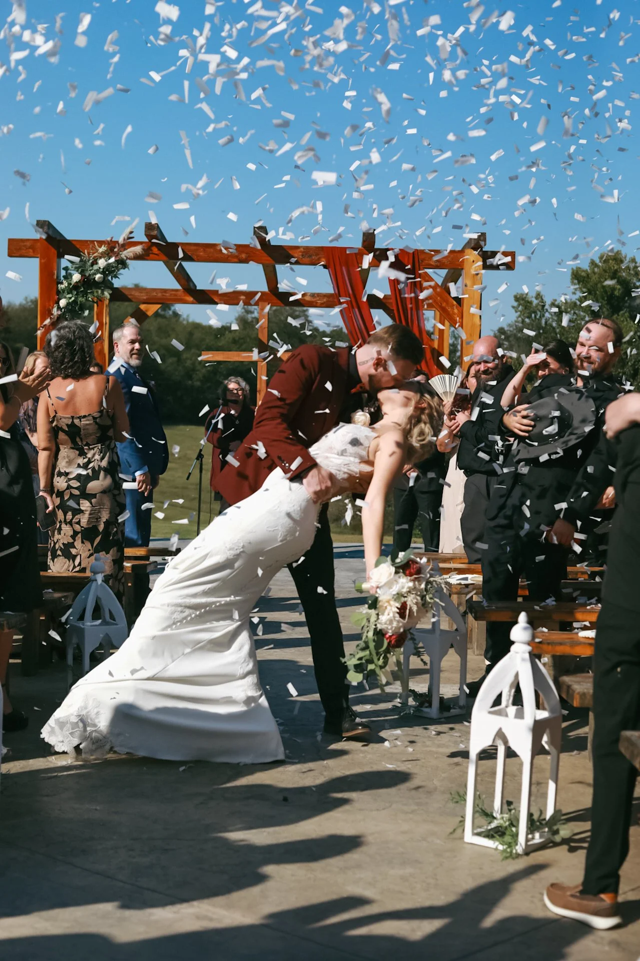 Wedding ceremony with confetti, bride and groom kissing under a wooden arch, surrounded by guests in festive attire.