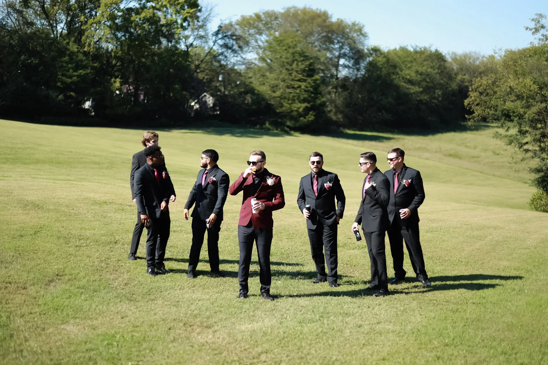 Group of men in formal attire standing on grass, wearing sunglasses and holding beverages, with lush greenery in the background.
