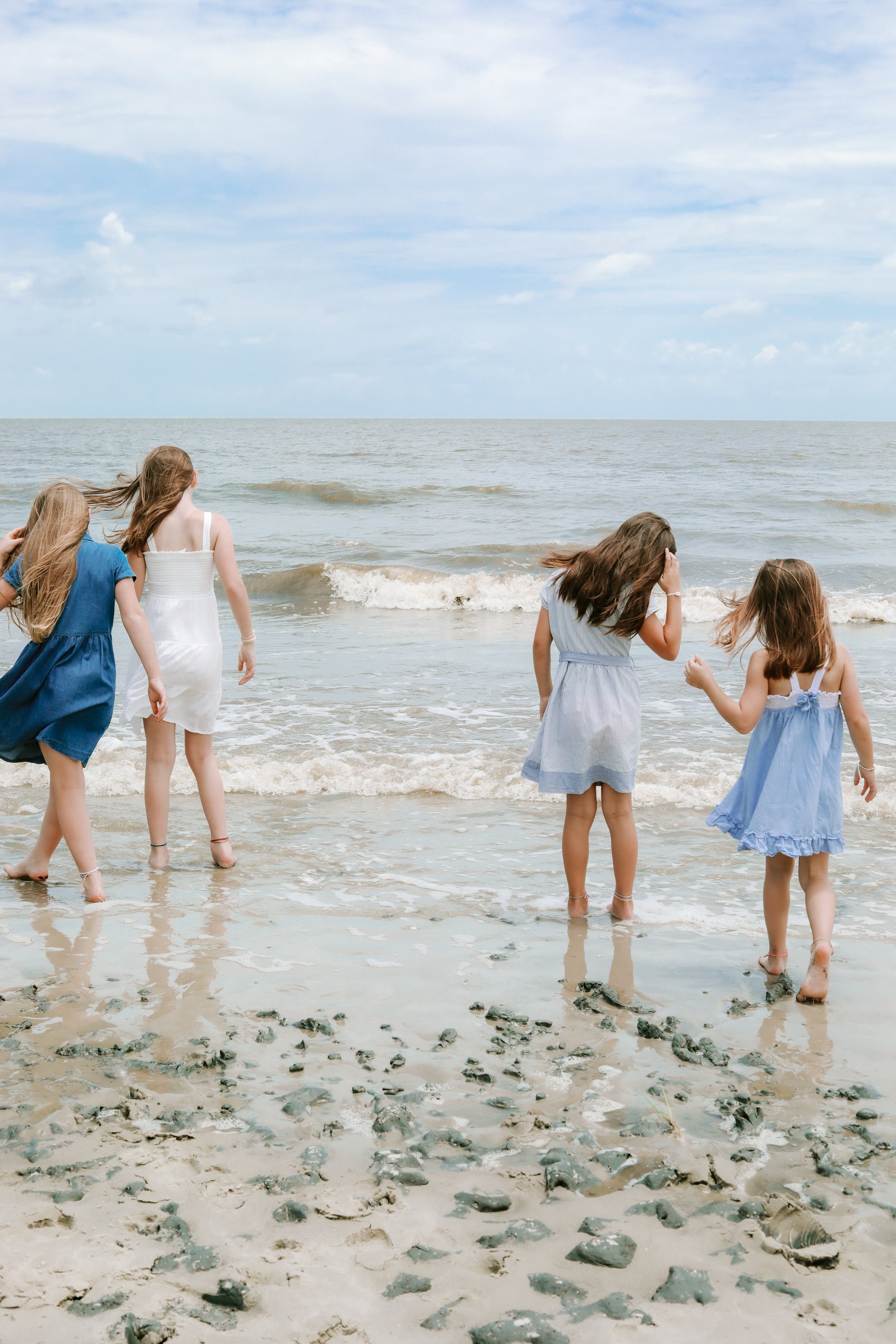 Group of girls in dresses standing by the ocean shore, with waves and cloudy sky.