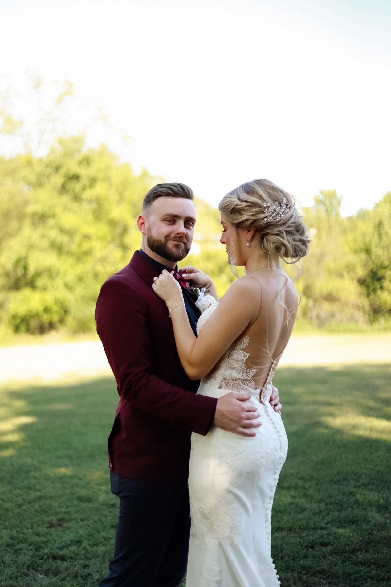 Bride and groom standing together outdoors in a grassy area, with the groom wearing a burgundy suit and the bride in a white lace wedding dress.