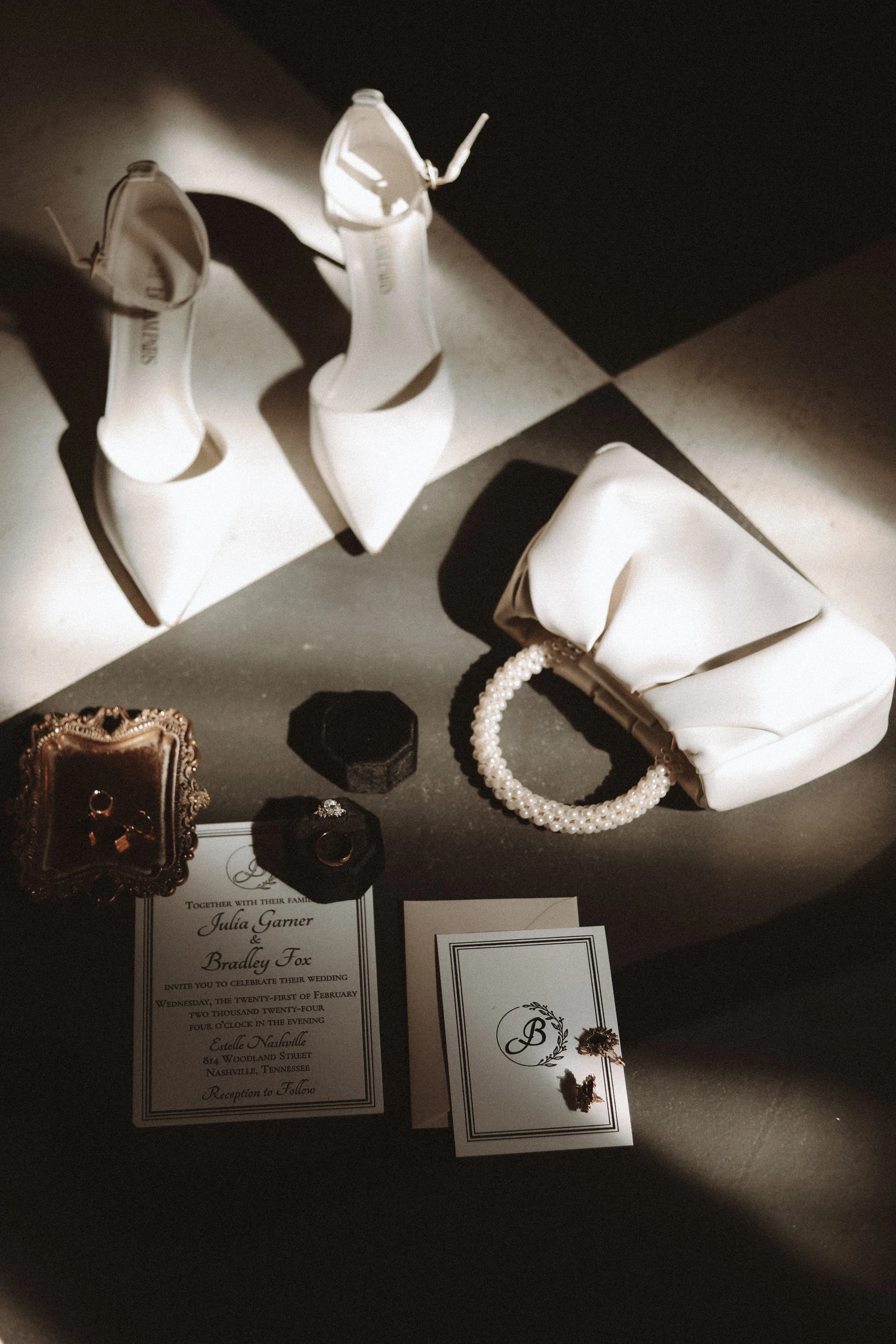 Wedding flat lay featuring white high heels, a pearl-handled clutch, an invitation, gold rings, a jeweled ring in a box, and dried flowers on a dark surface.