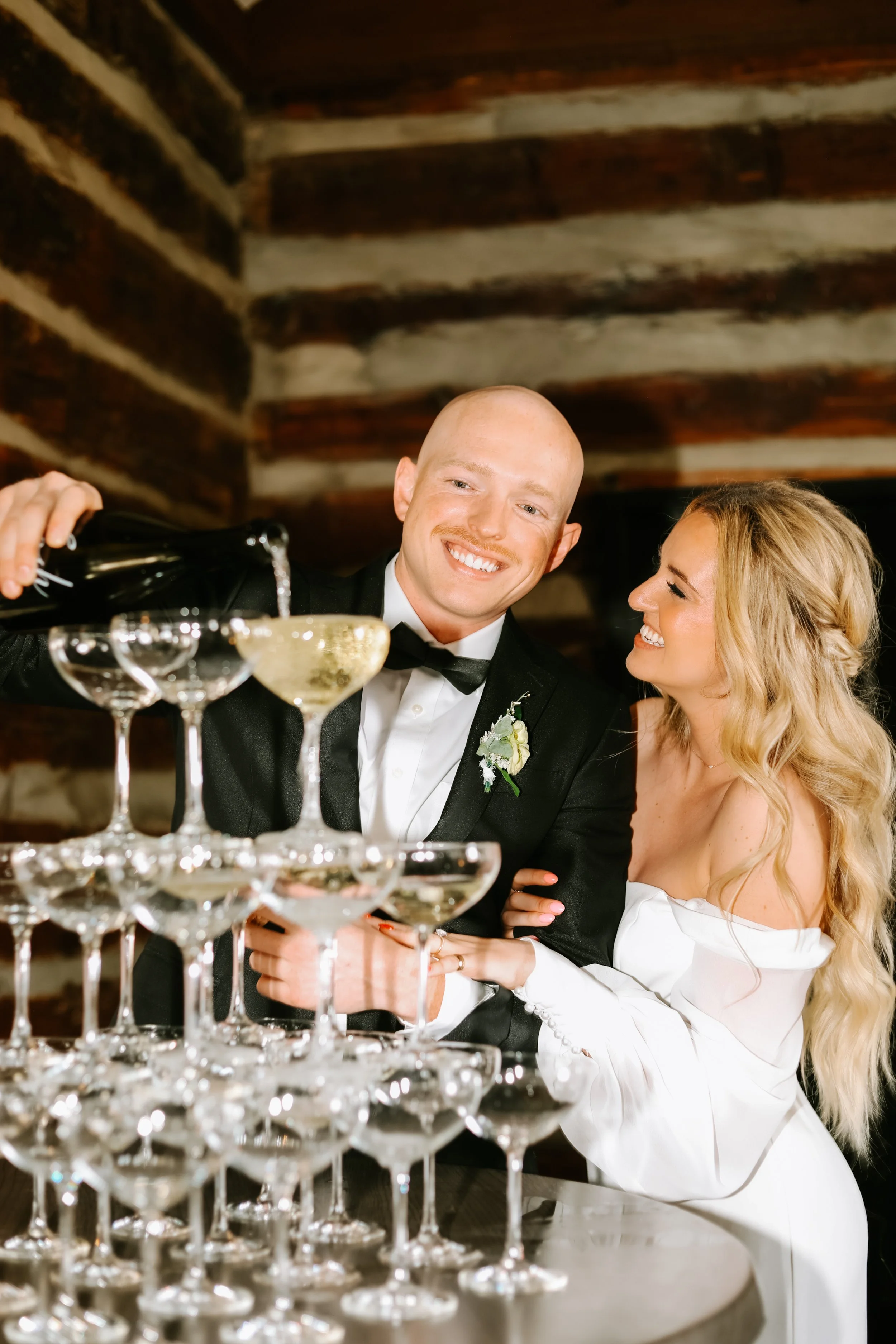A couple celebrating, with a man pouring champagne into a tower of glasses while the woman smiles and embraces him.