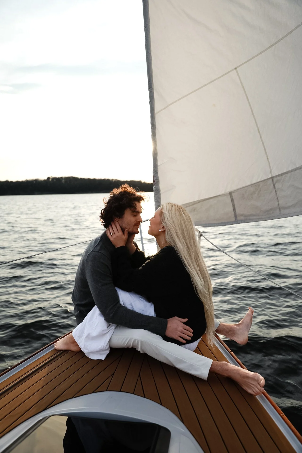 A couple sitting closely and embracing on the deck of a sailboat with a large sail, surrounded by water and distant shoreline under a clear sky.