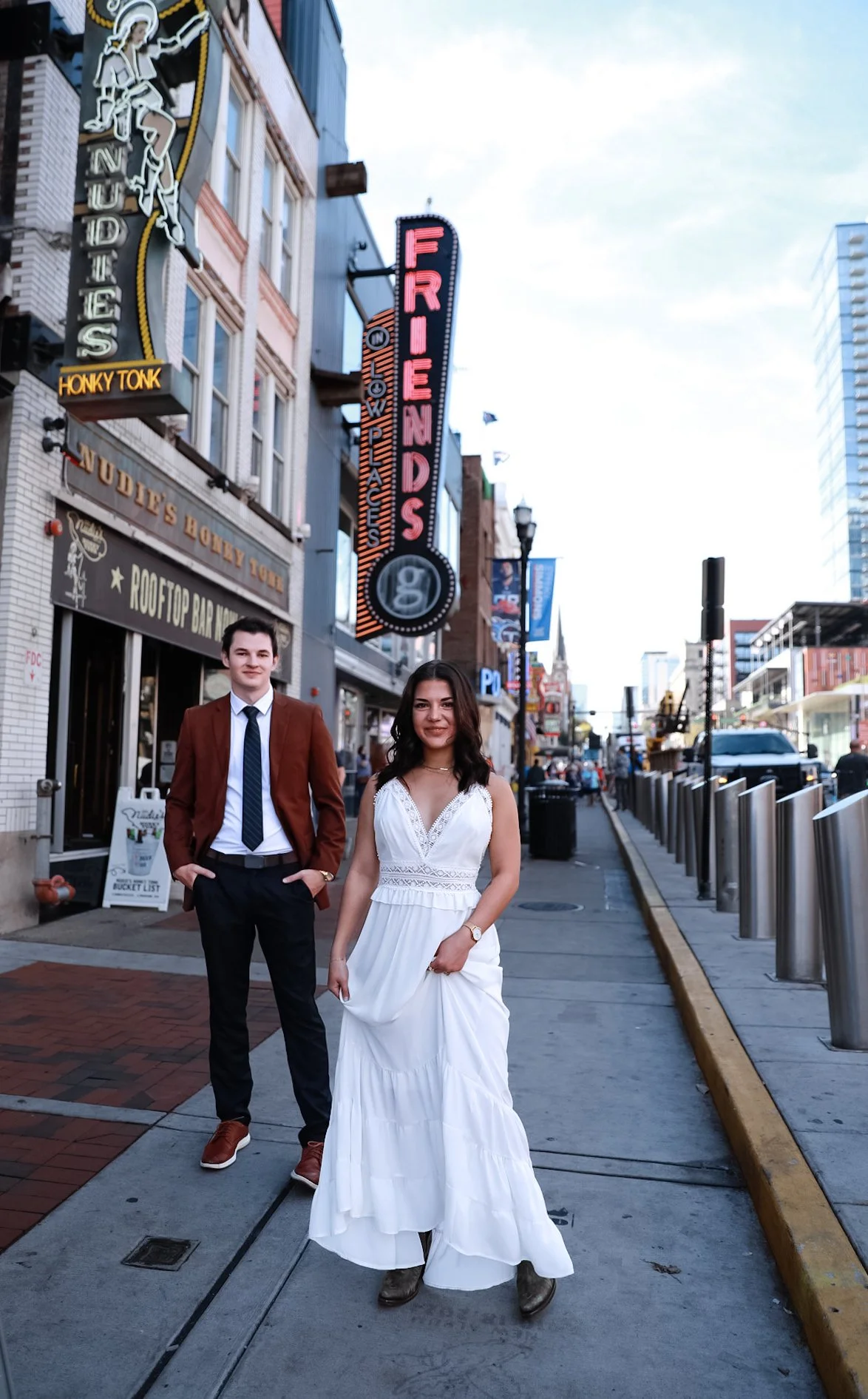 A man in a brown jacket and black pants stands next to a woman in a white dress on a city street, with neon signs for bars in the background, including a sign for "Nudie's Honky Tonk."