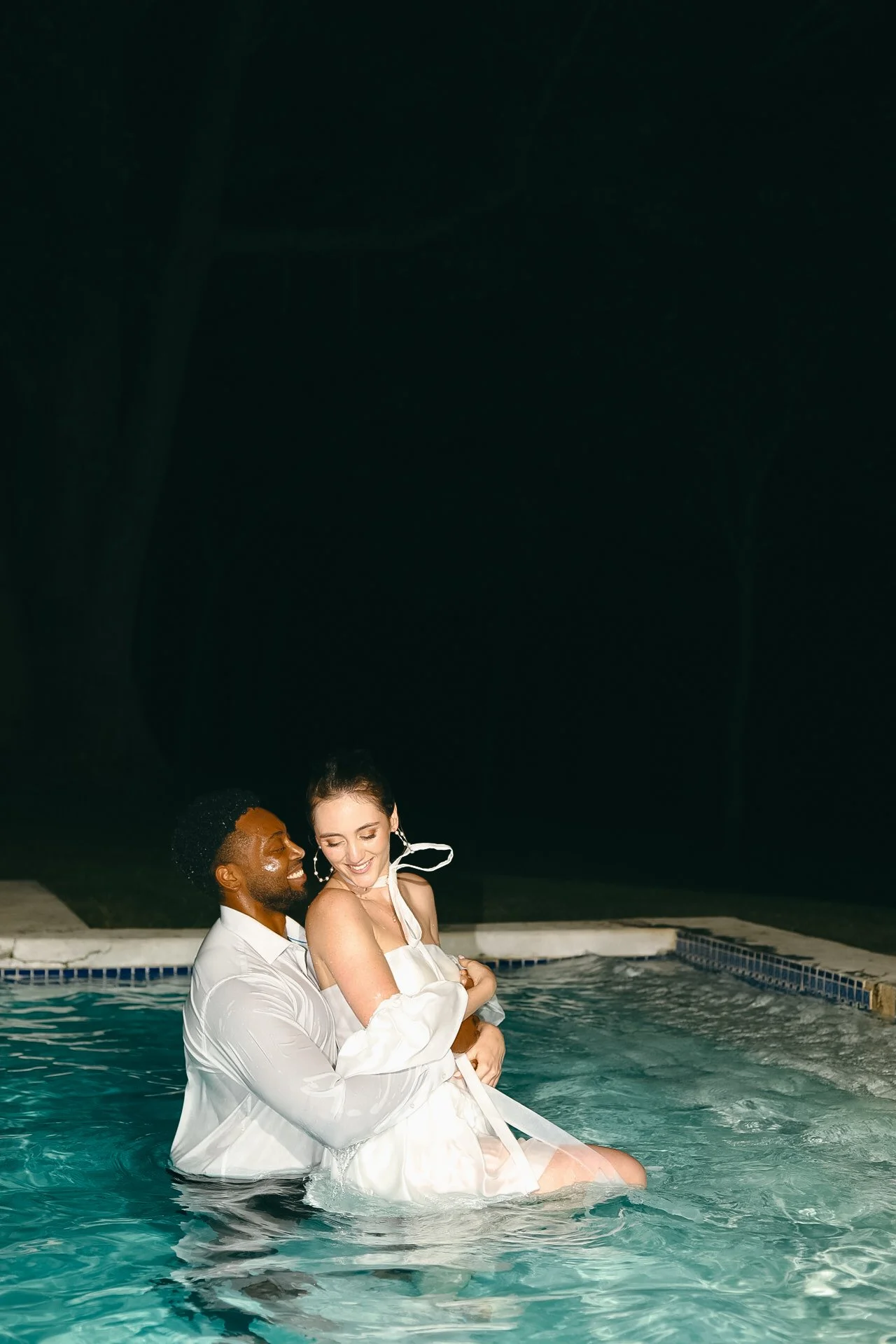 A man and woman in formal attire embracing in a swimming pool at night.