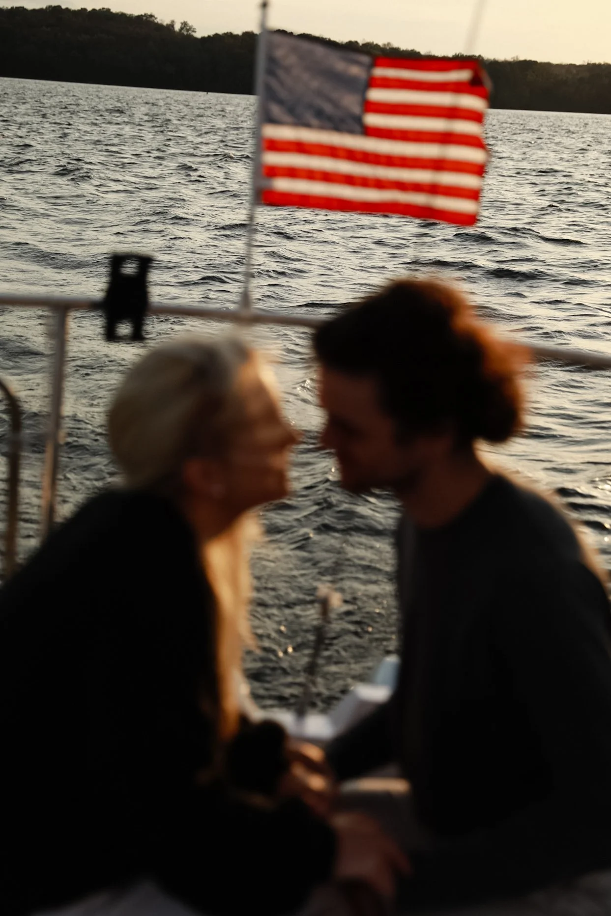 Blurry couple sitting on a boat with an American flag in the background and a view of the water.