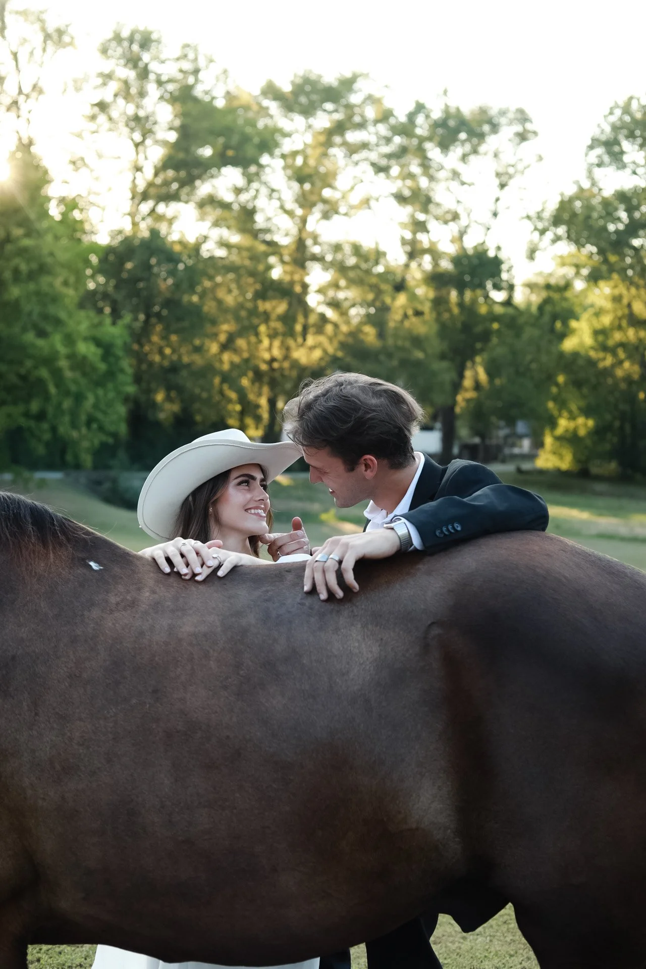 A young couple in formal attire, with the woman wearing a white wide-brimmed hat, smiling and leaning on a horse's back, outdoors in a park with trees and sunlight.