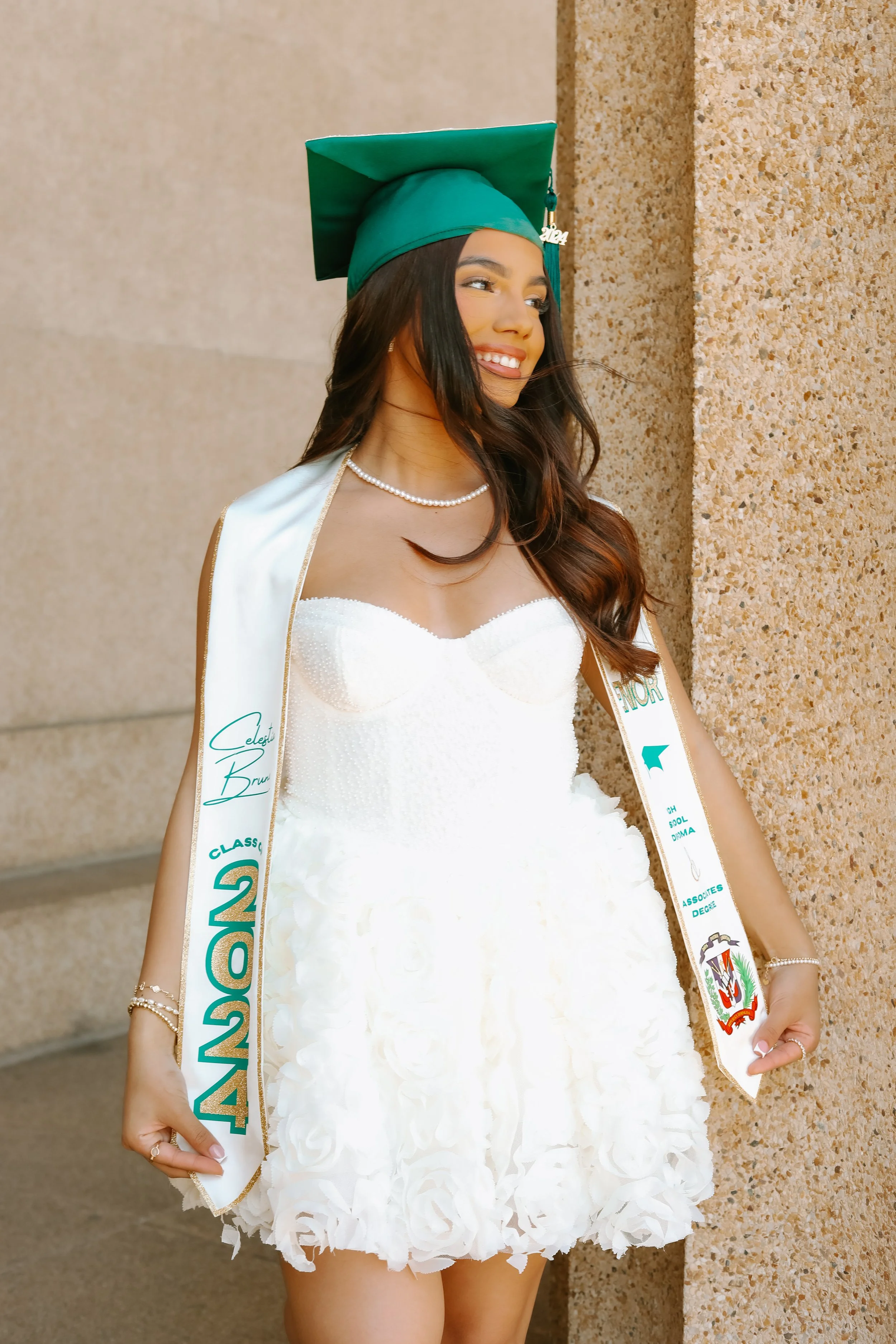 Young woman in a white dress wearing a green graduation cap and a sash with "Class of 2024," standing against a stone wall, smiling and looking away.