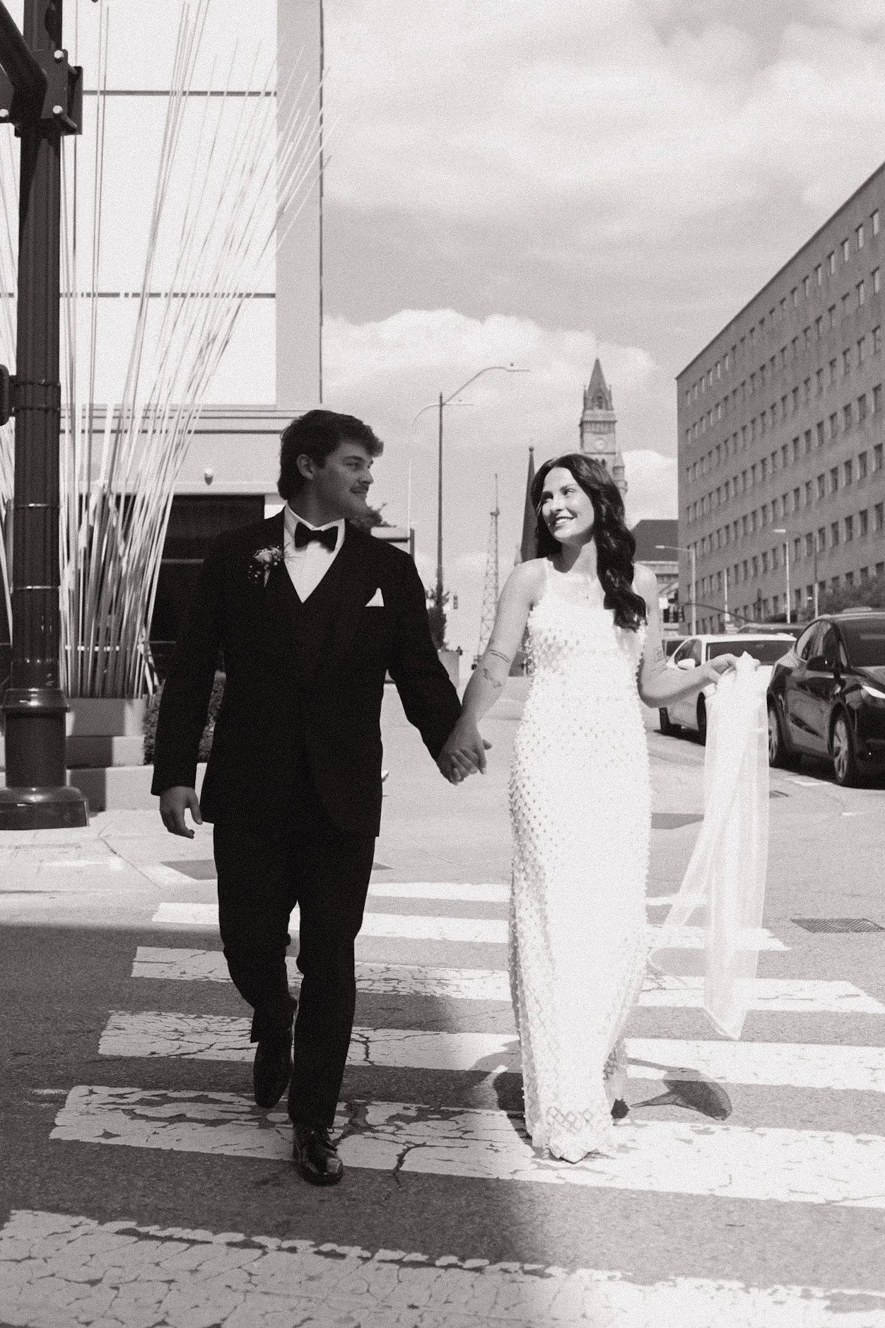 A black and white photo of a newlywed couple walking hand in hand across a crosswalk on a city street. The groom is wearing a black tuxedo, and the bride is in a white wedding dress holding a veil.