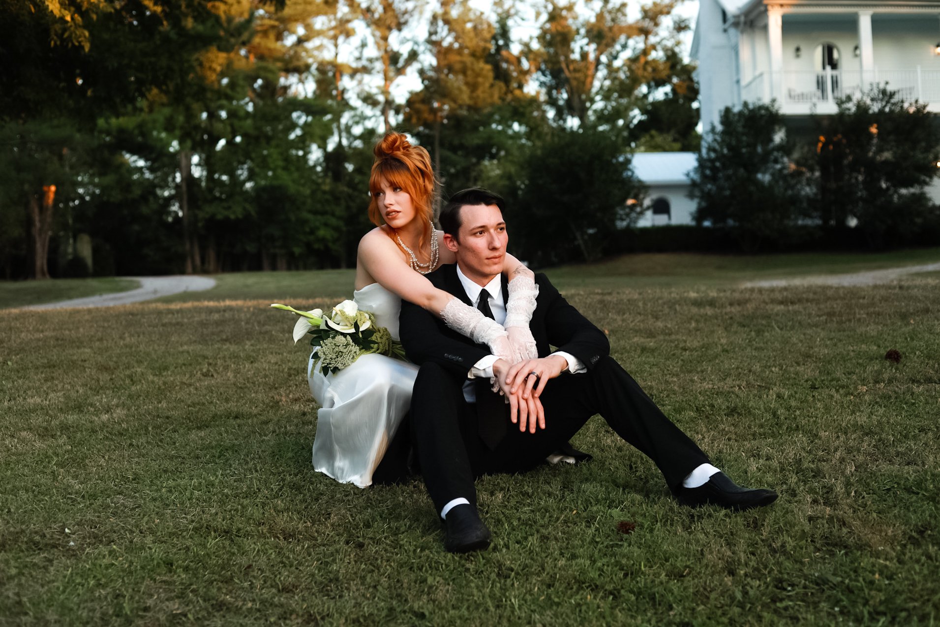 A newlywed couple sitting on the grass in a park, with the bride embracing the groom from behind. The bride has red hair styled in an updo, wears a white wedding dress, and holds a bouquet of white flowers. The groom has dark hair, wears a black tuxedo with a white shirt and black tie, and is sitting with his legs extended forward and leaning slightly to one side.