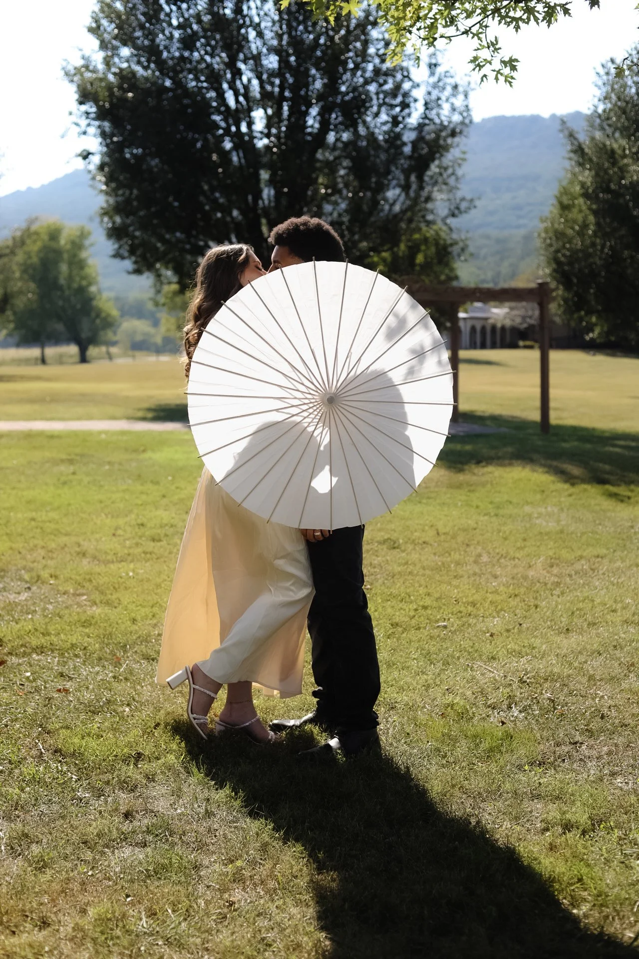 A couple standing on grass, kissing behind a large white parasol, with trees and mountains in the background.