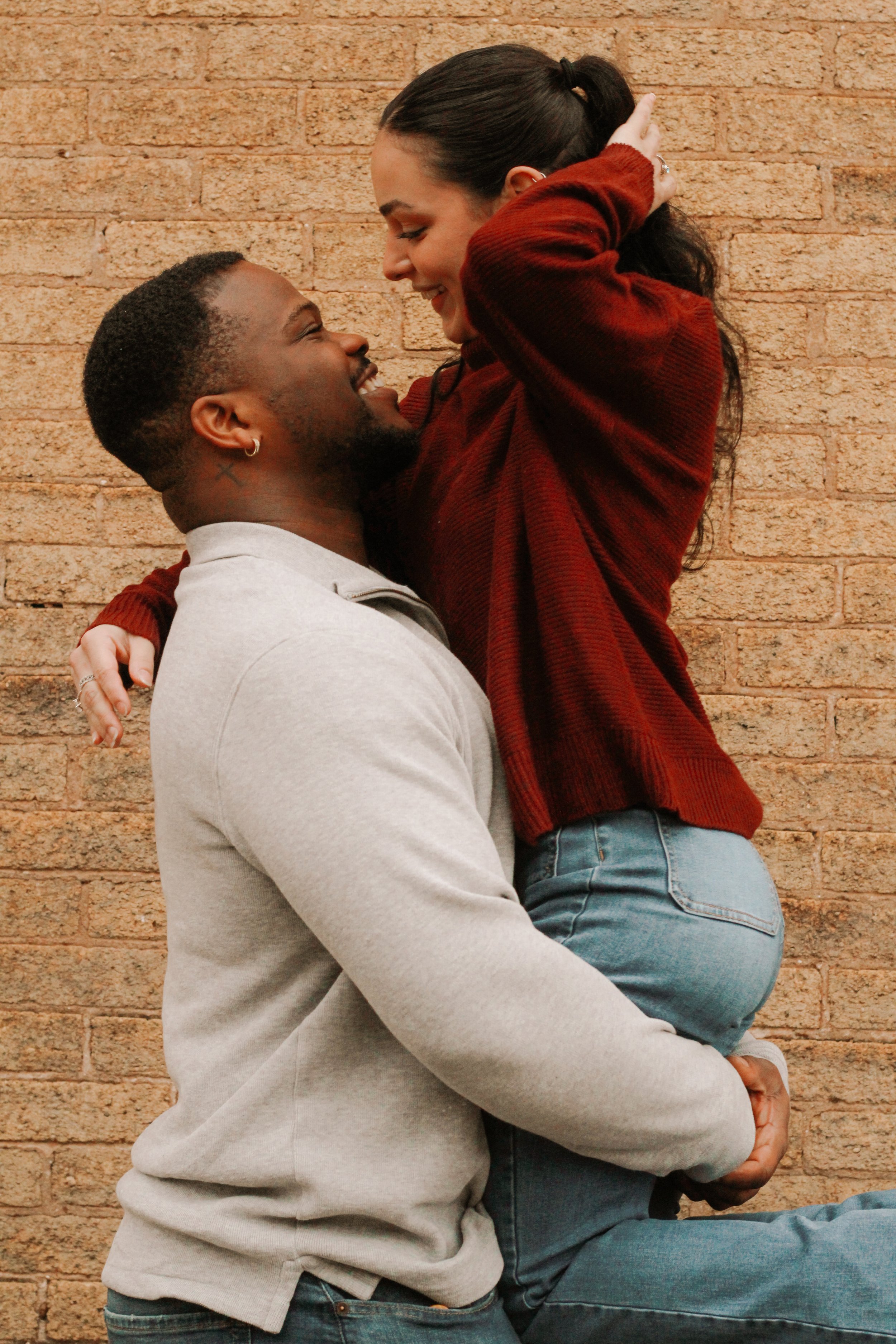 A couple embracing affectionately, with the man lifting the woman, in front of a brick wall. The woman is wearing a red sweater and jeans, and the man is wearing a gray sweater.