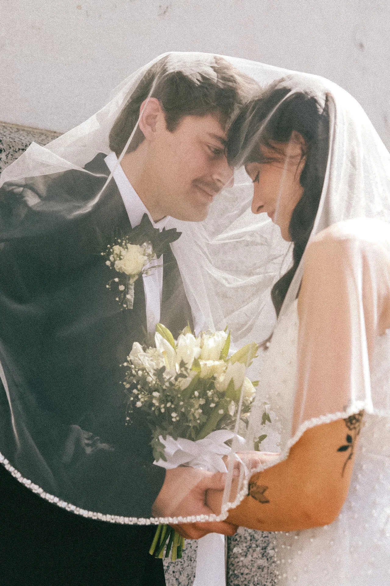 A bride and groom under a wedding veil, holding a bouquet of white flowers, with foreheads touching
