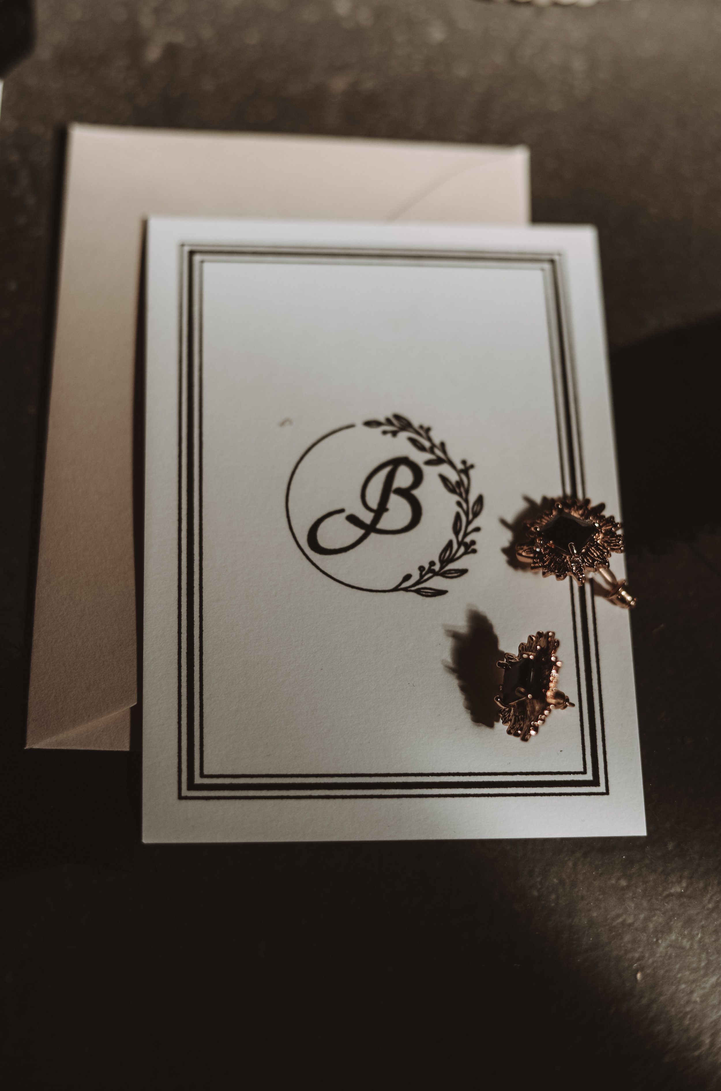 Elegant card with an embossed letter B and intricate border design next to two vintage earrings.