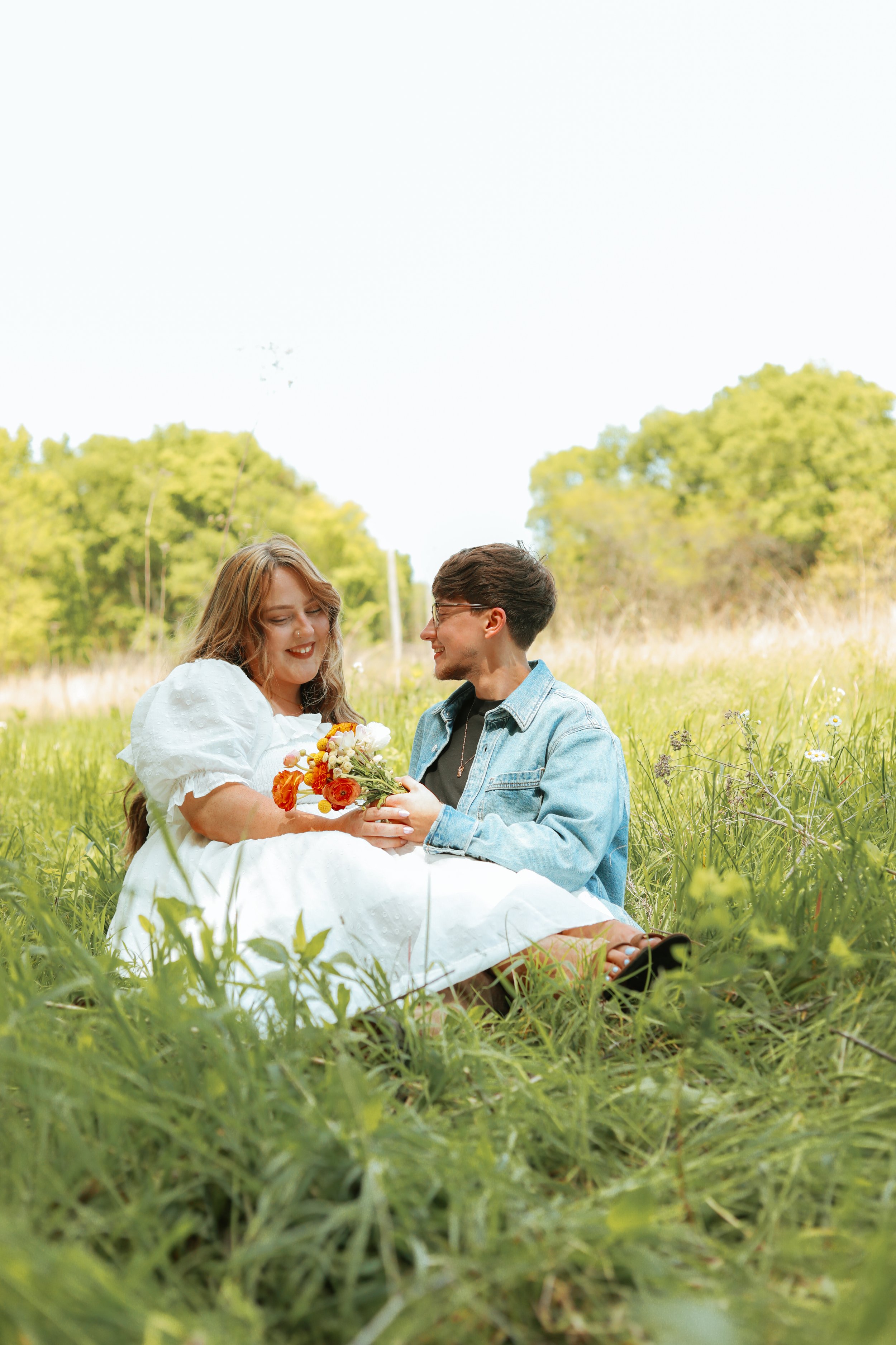 Two people sitting on grass in a park, sharing a bouquet of flowers, with trees in the background.