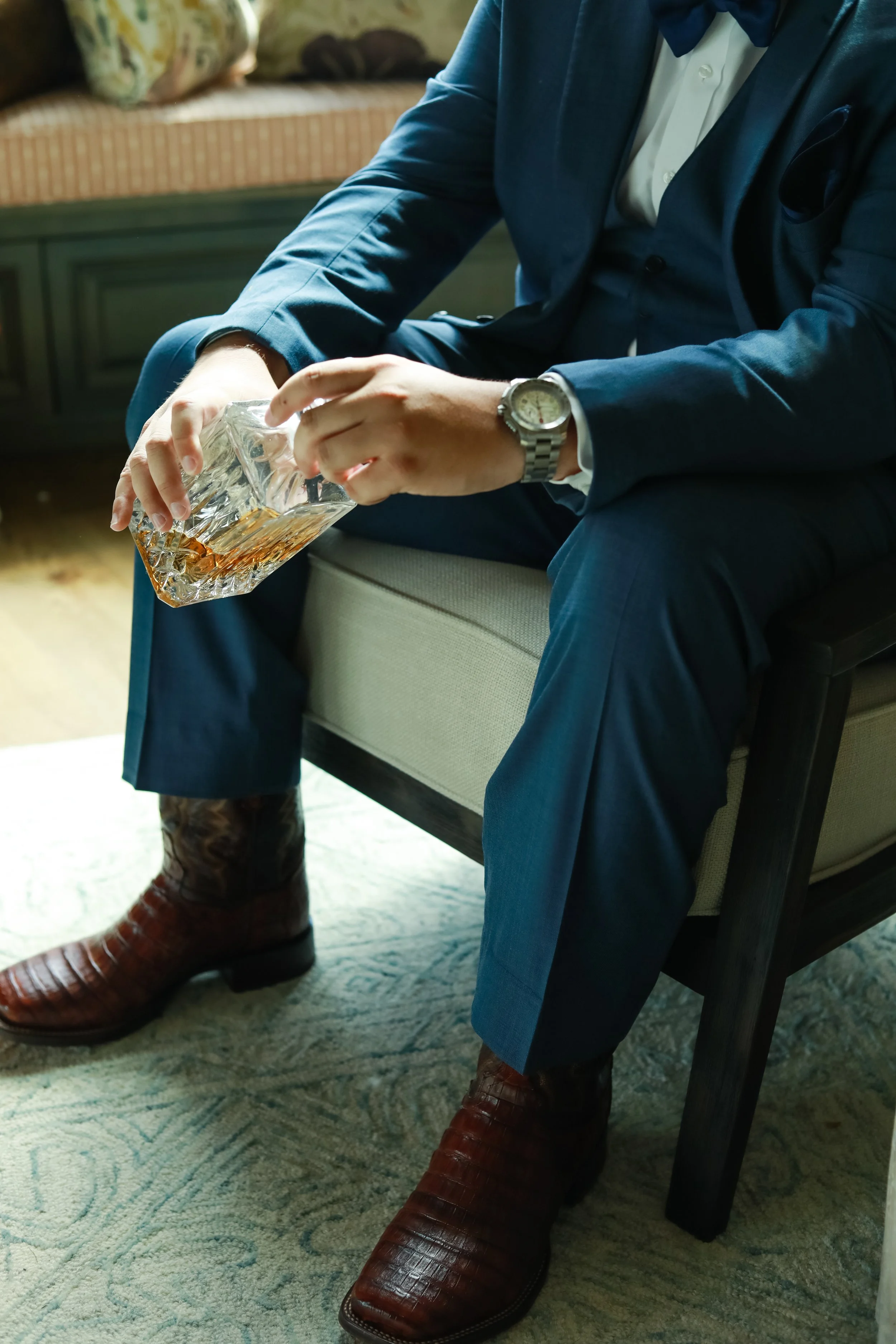 Man in blue suit sitting on a chair, holding a glass decanter with amber liquid, wearing brown leather shoes, and a watch.