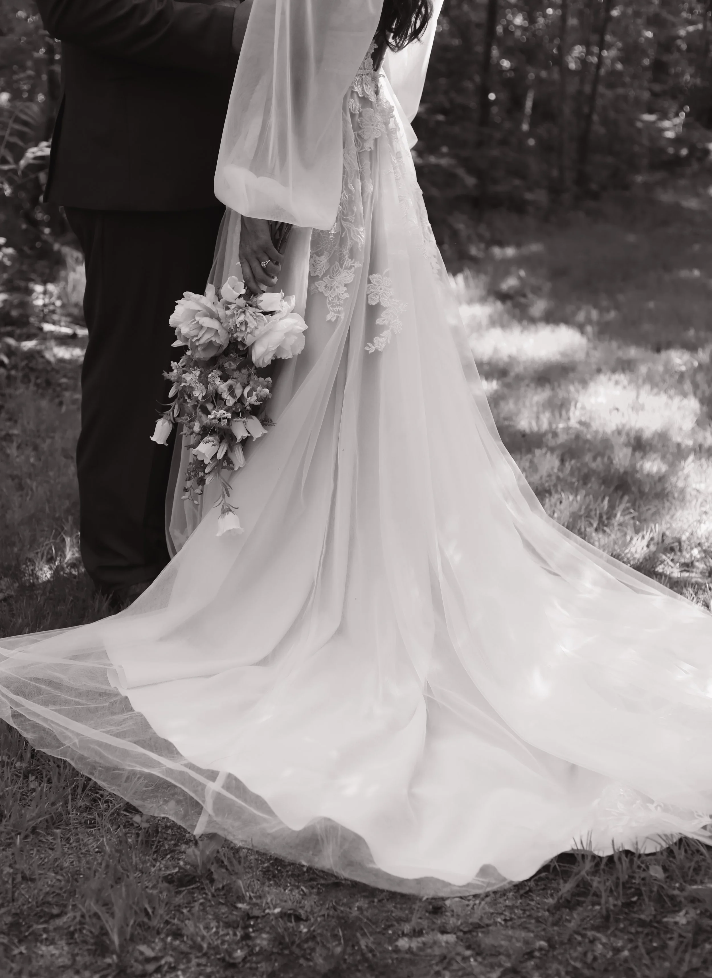 Black and white photo of a bride in a flowing wedding dress holding a bouquet, standing next to a groom in a suit in an outdoor setting.