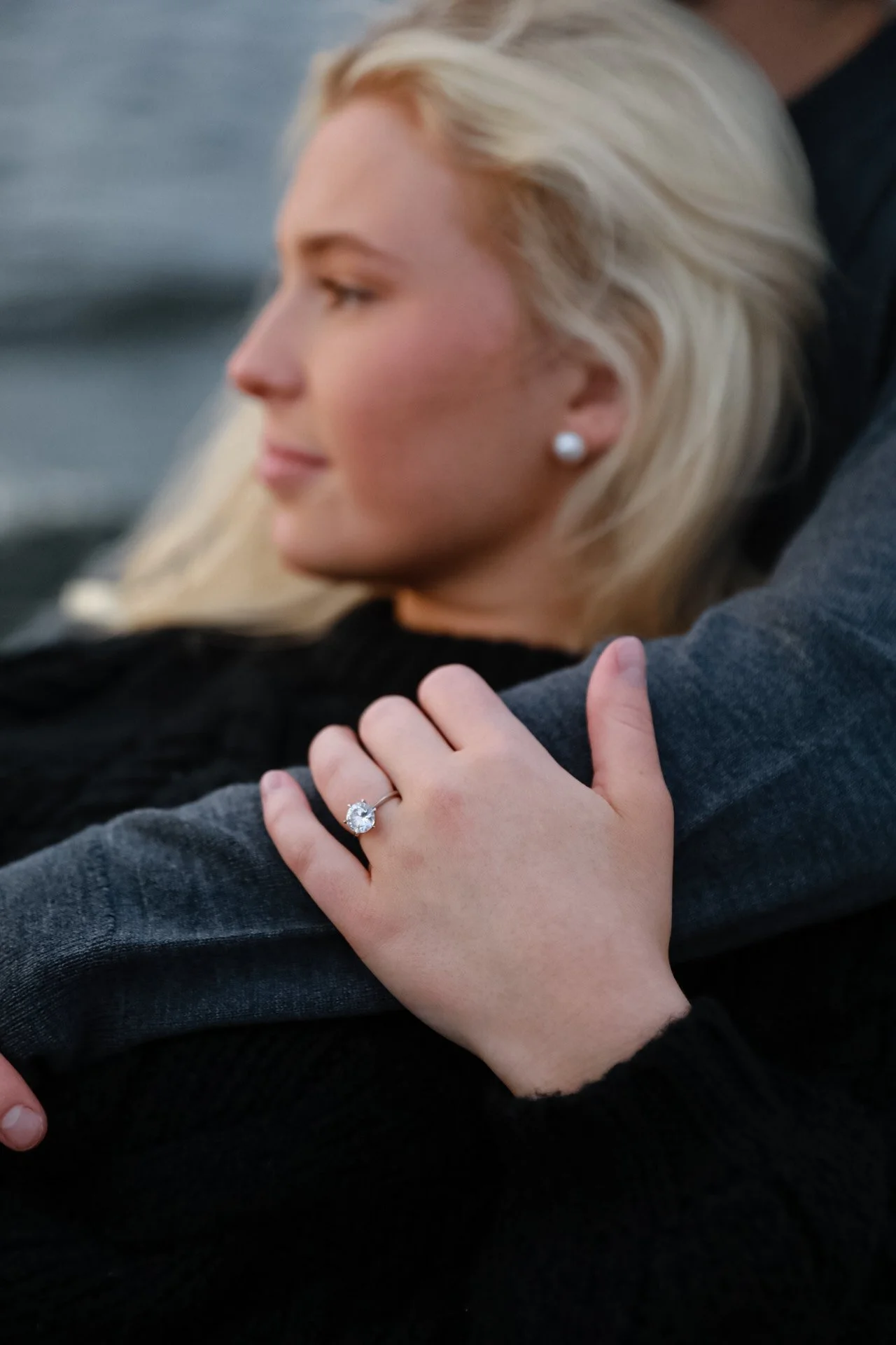 Close-up of a woman with blonde hair wearing pearl earrings and displaying a diamond ring on her hand.
