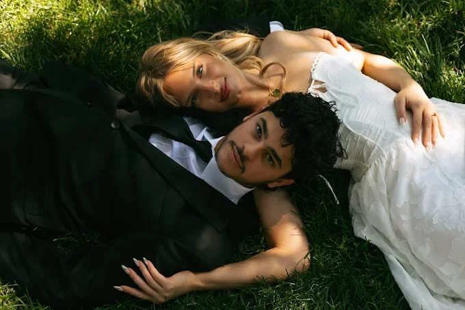 A newlywed couple sitting on the grass in a park, with the bride embracing the groom from behind. The bride has red hair styled in an updo, wears a white wedding dress, and holds a bouquet of white flowers. The groom has dark hair, wears a black tuxedo with a white shirt and black tie, and is sitting with his legs extended forward and leaning slightly to one side.