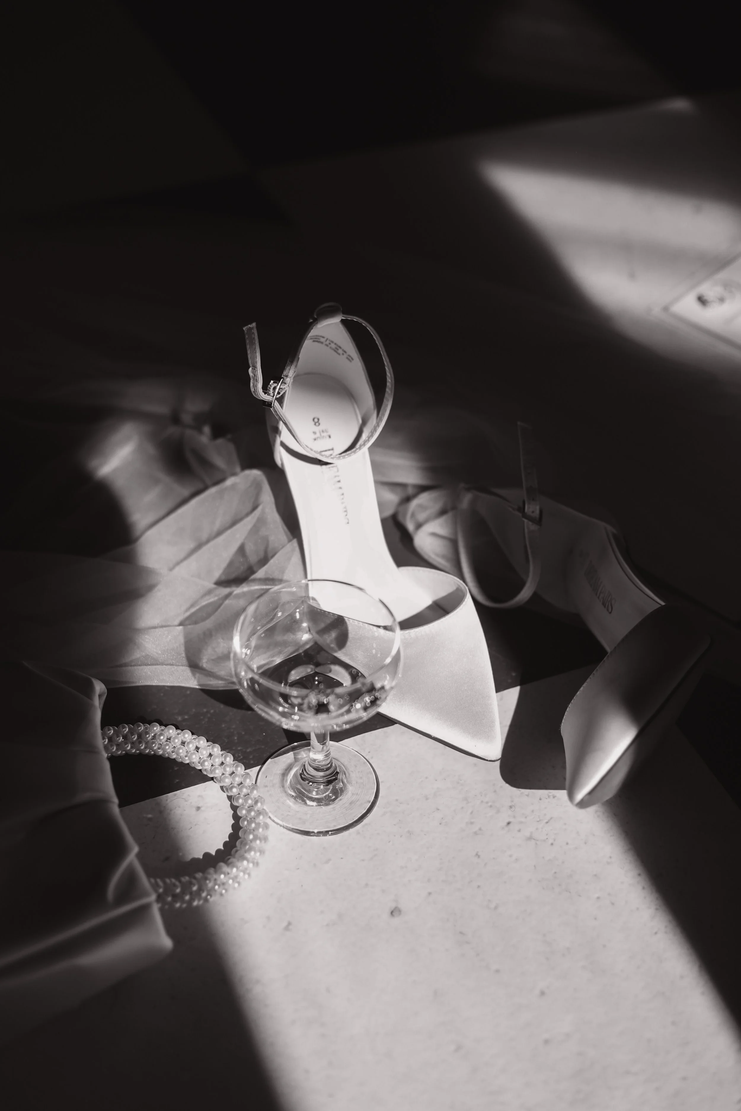 Black and white image of high heels, champagne glass, and a beaded necklace on a textured surface, with dramatic lighting.