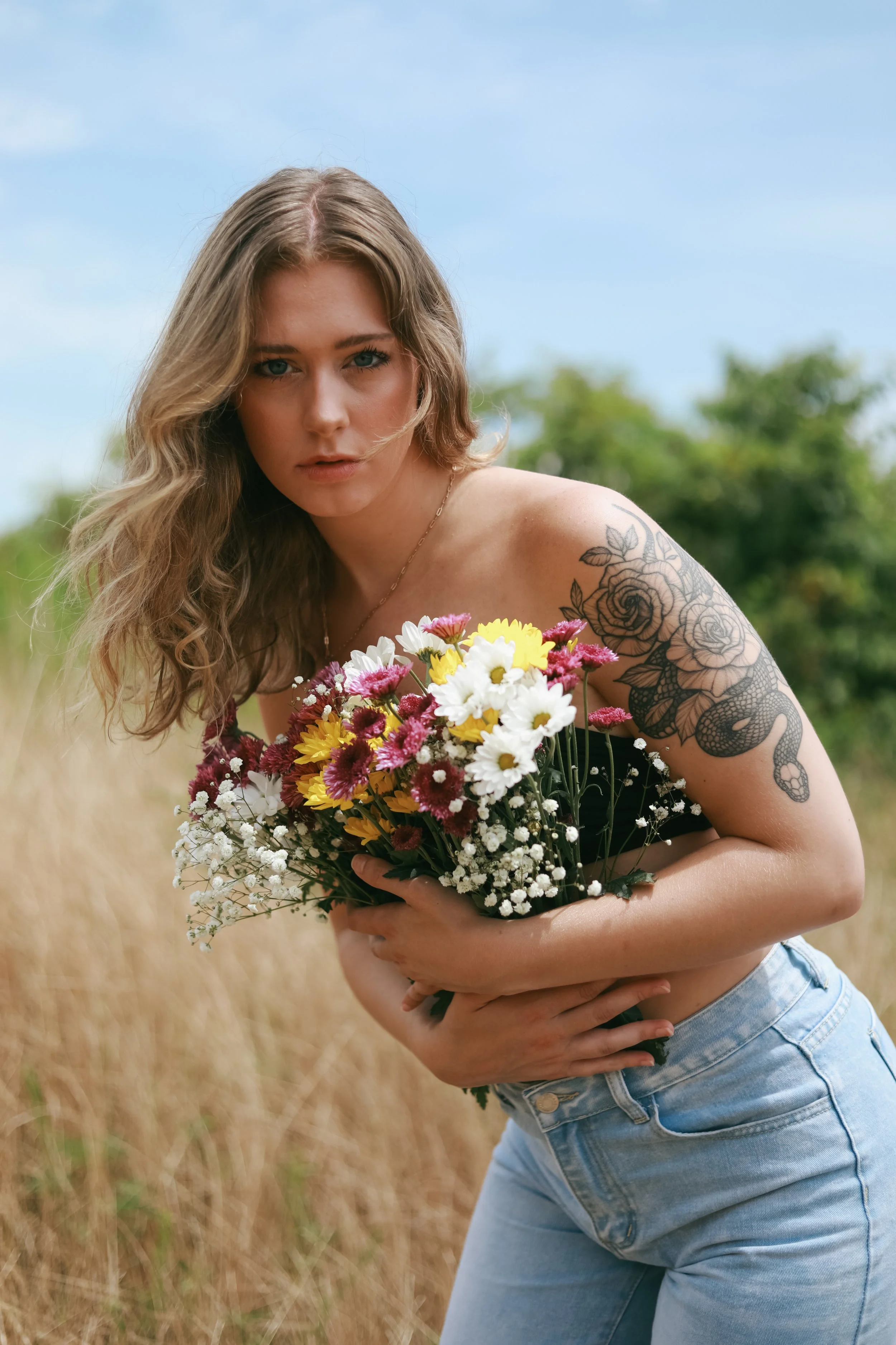 Woman holding a bouquet of mixed flowers with a tattoo on her arm in an outdoor setting.