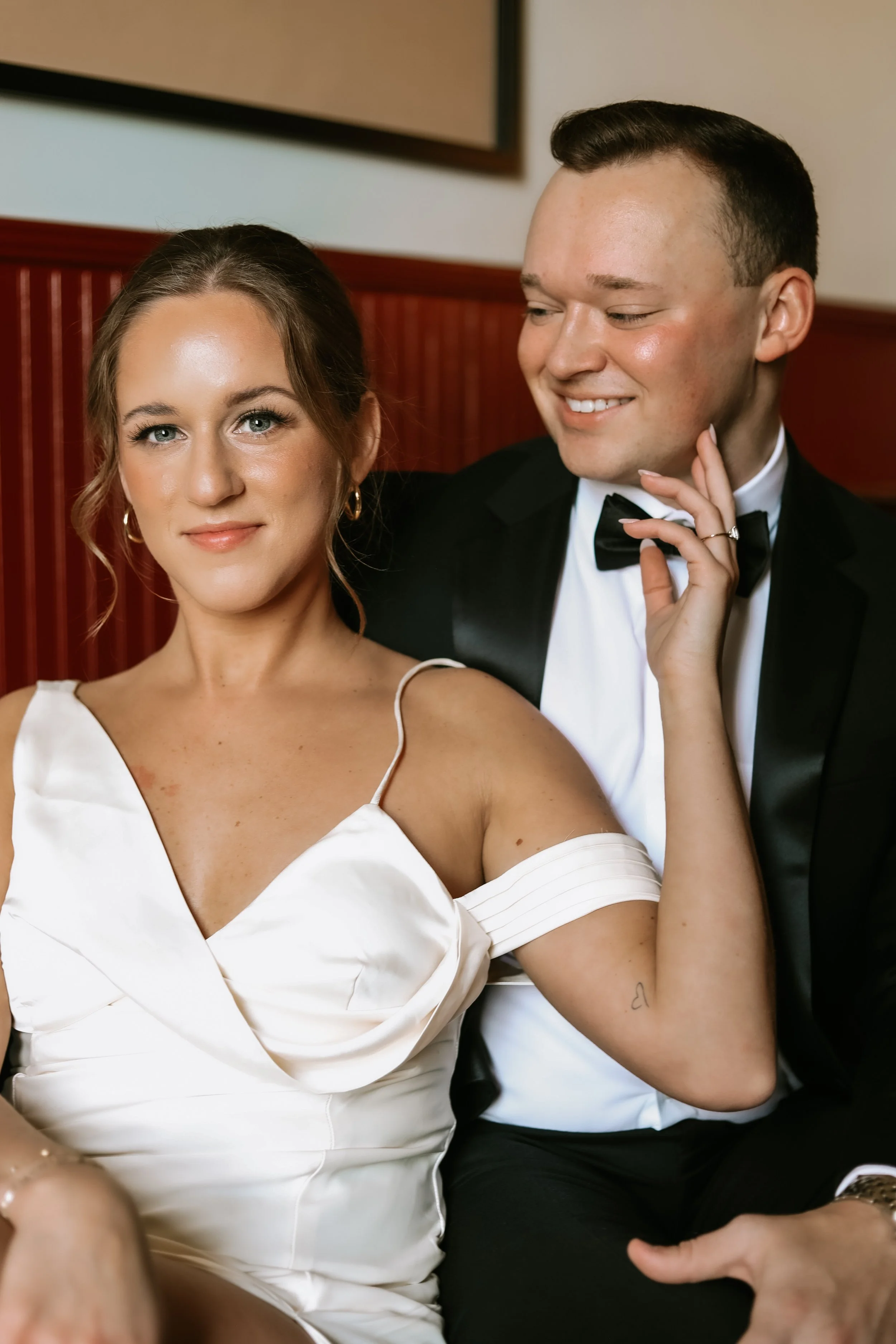 Elegant couple posing together, woman in white dress and man in black tuxedo with bow tie, sitting in a decorated interior with a red wooden wall.