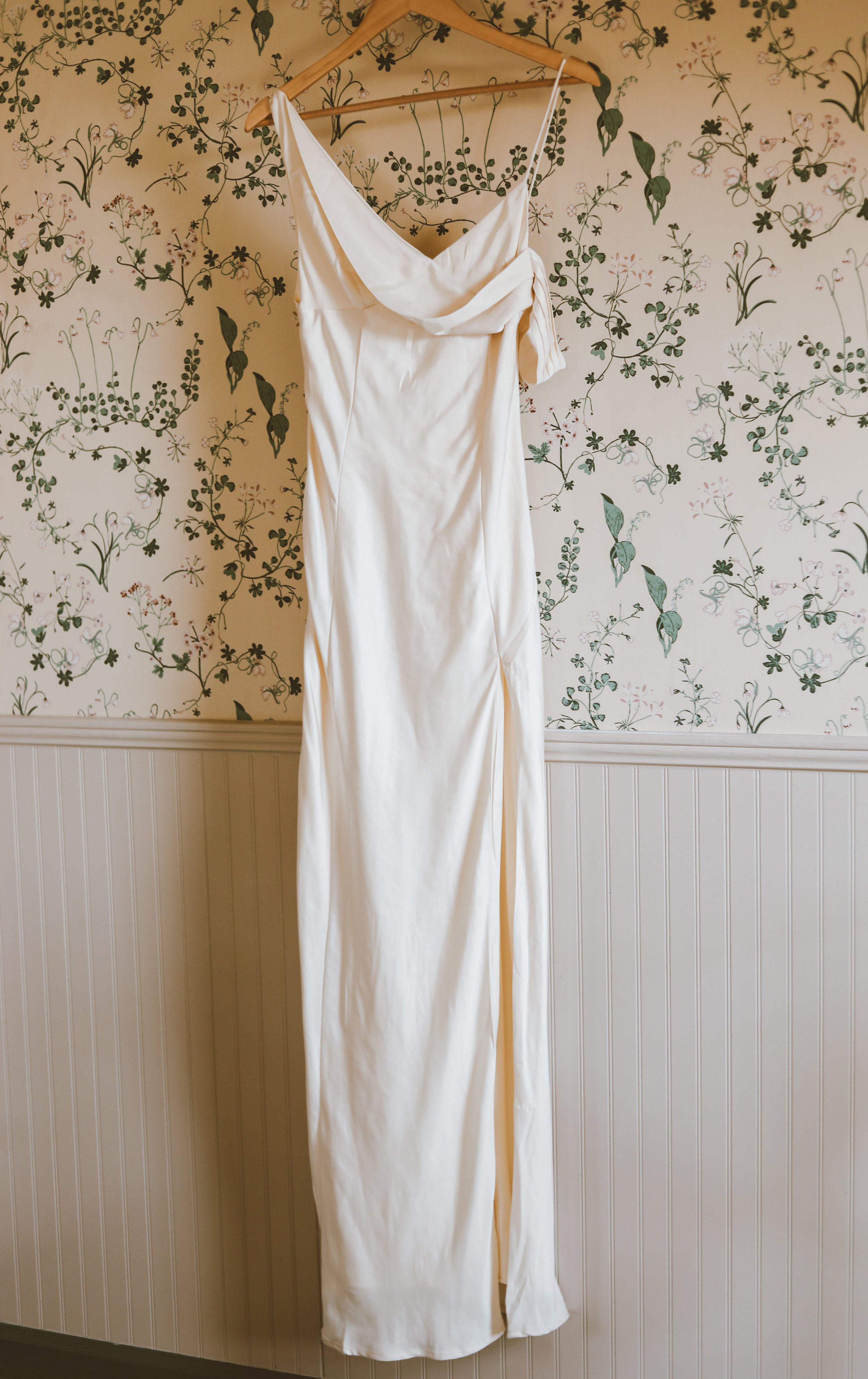 A white sleeveless dress with a draped neckline hanging on a wooden hanger against floral wallpaper.