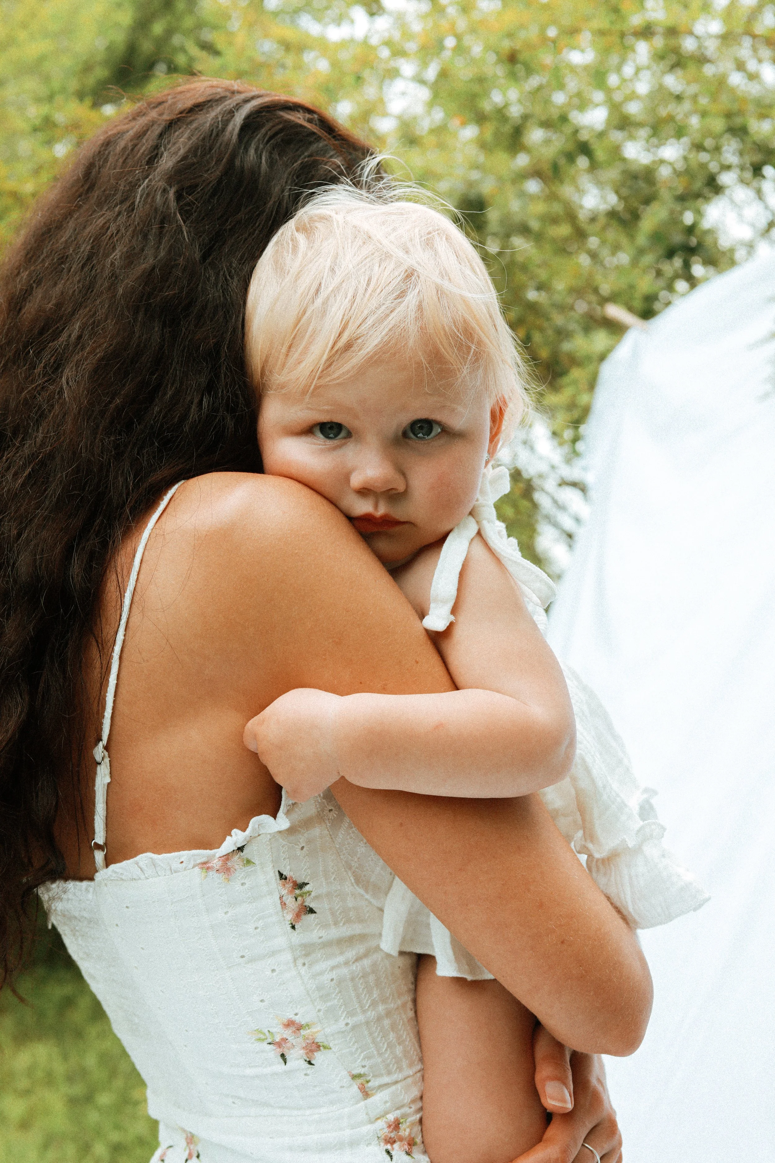 A woman with long hair holding a baby outdoors in a garden. The baby is wearing a white outfit and looks at the camera.