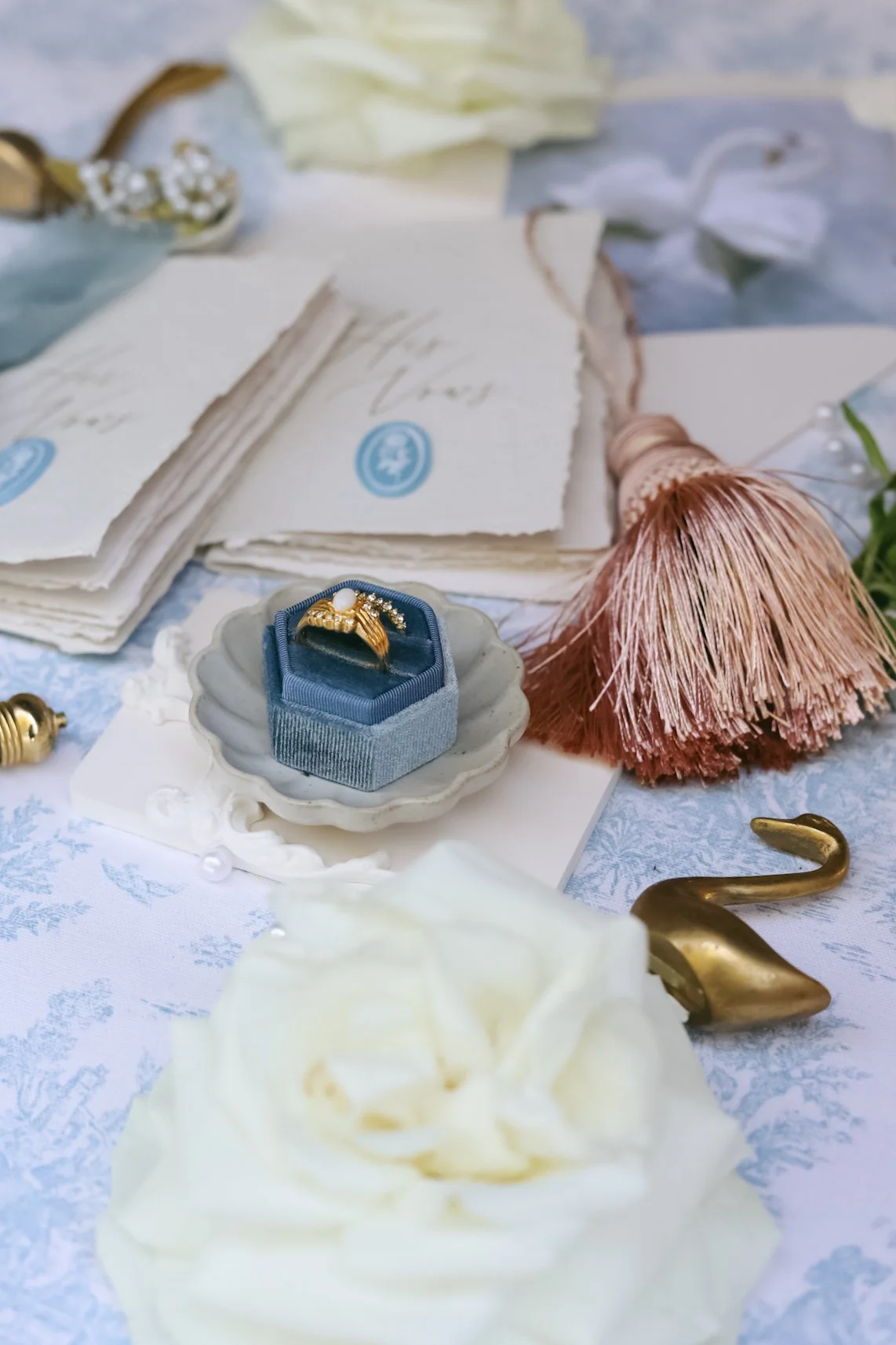 Wedding-themed flat lay featuring a gold ring with pearls in a blue velvet box, surrounded by white roses, wedding vows booklets, and decorative tassel on a patterned tablecloth.