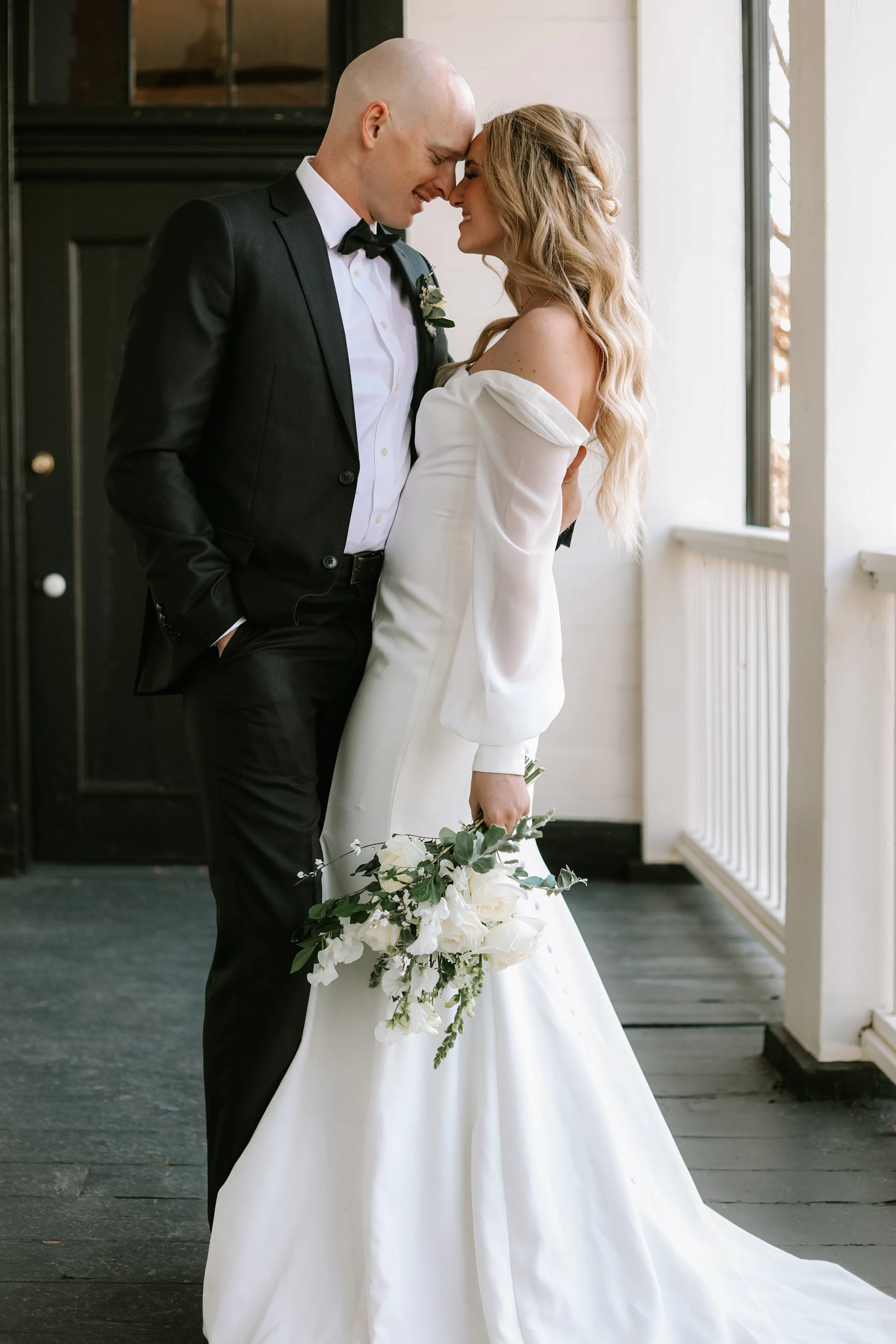 Bride and groom embracing on a porch, bride holding a bouquet of white flowers, wearing a white gown, groom in a black suit with a bow tie.