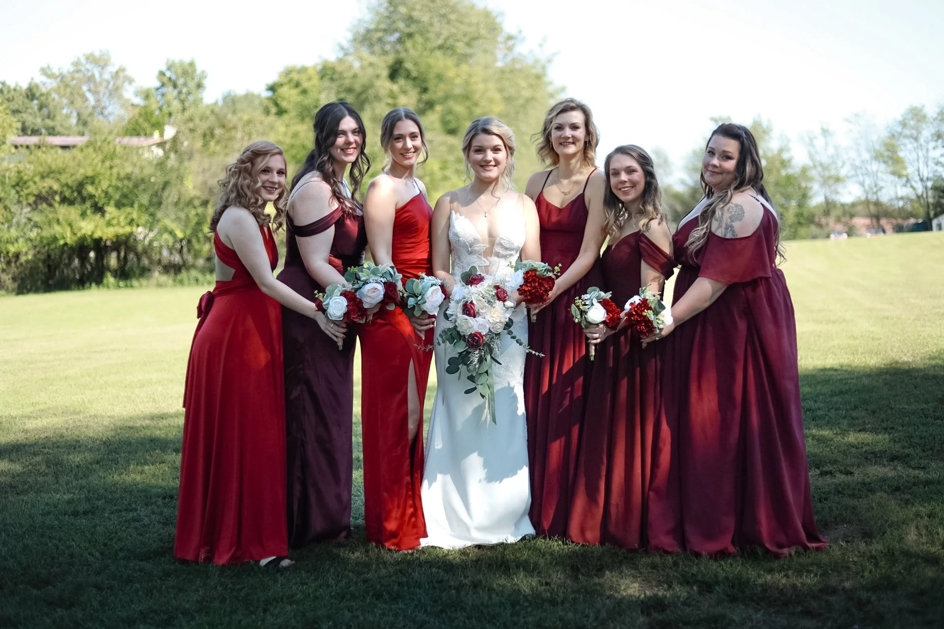 Bride with bridesmaids in red dresses holding bouquets on a lawn, surrounded by greenery.
