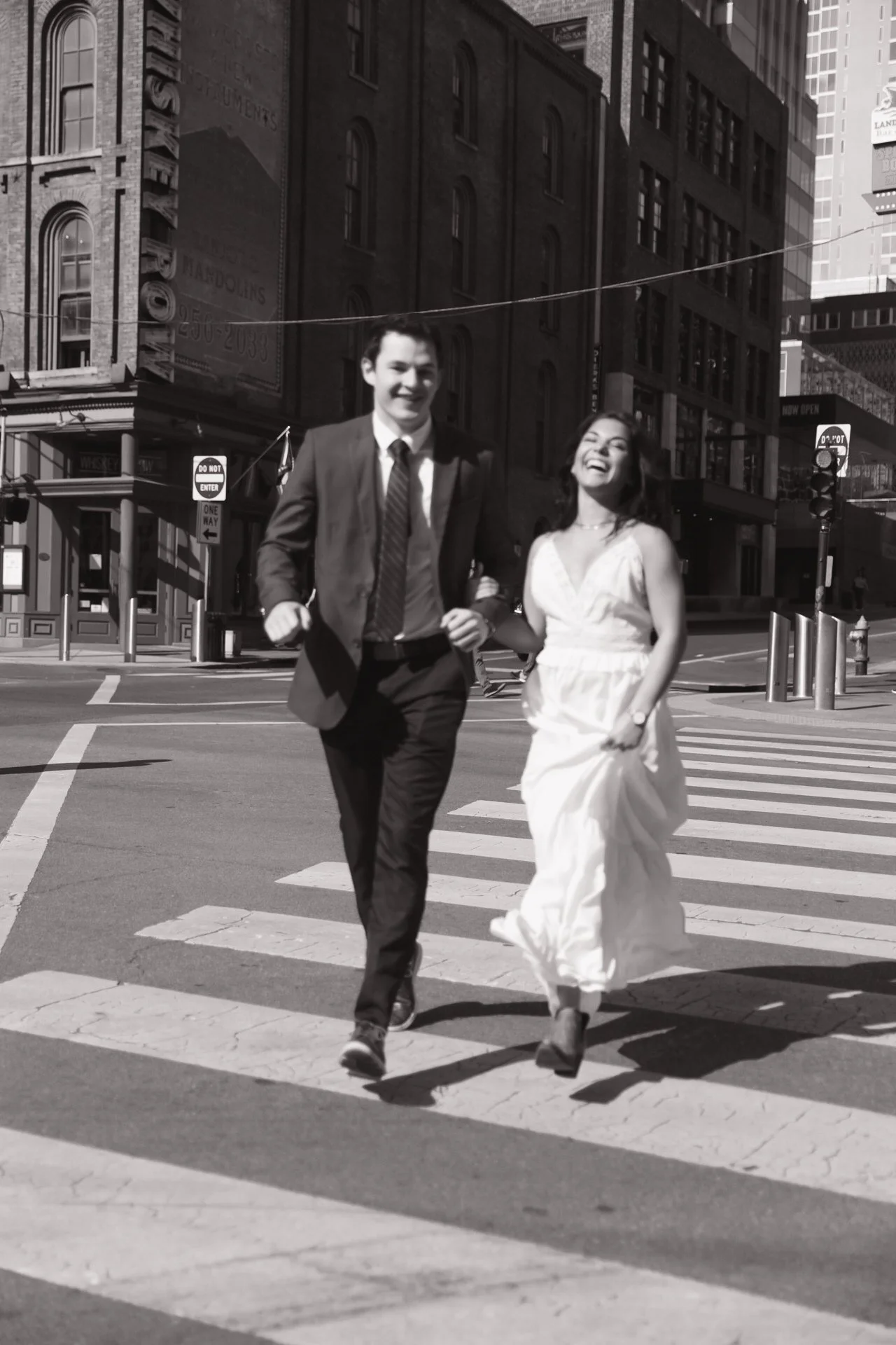Happy couple in formal attire walking across a city street crosswalk, with historic brick buildings in the background.