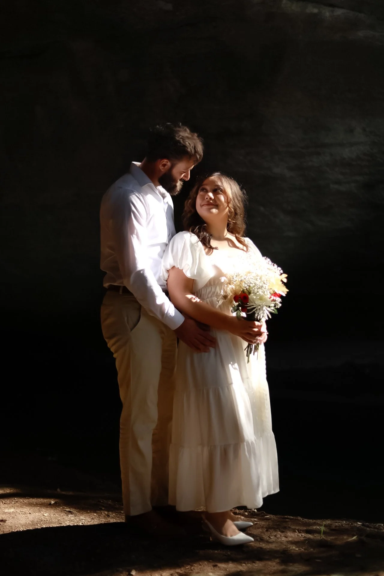 A couple in romantic attire stands in a dark setting, with the woman holding a bouquet and gazing up at the man, who is holding her waist. Sunlight softly illuminates them.