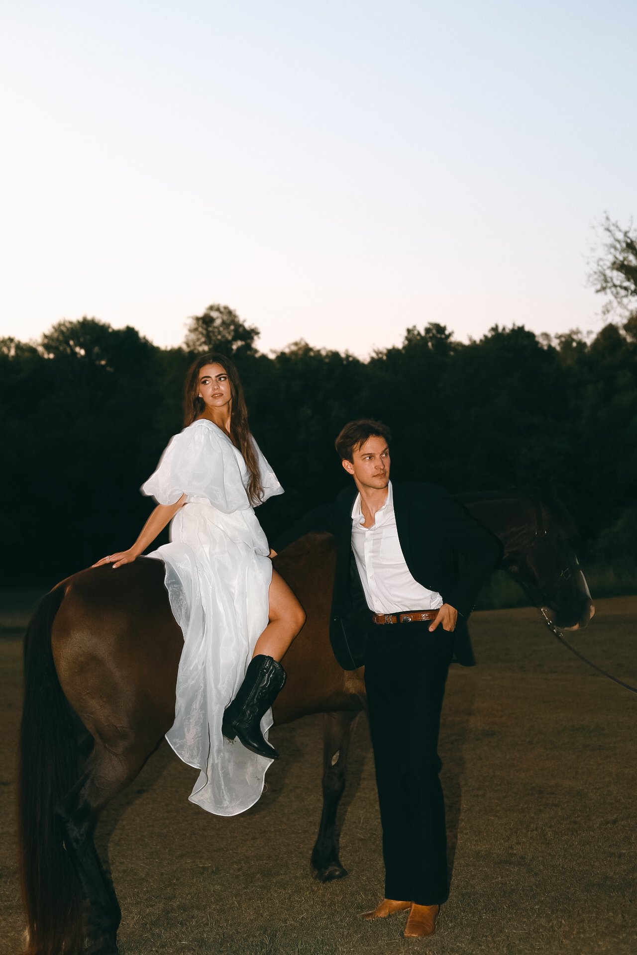 Woman in white dress sitting on a horse, man in black suit standing beside, outdoor setting.