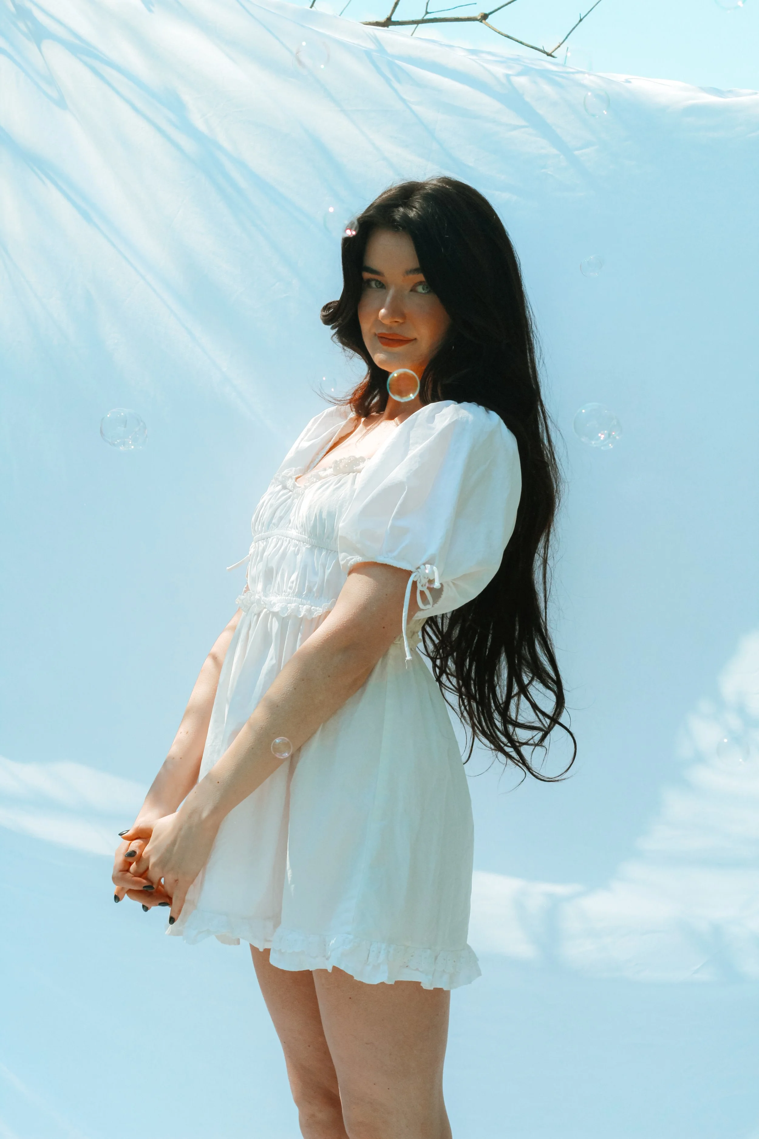 Woman in a white dress with long dark hair standing against a light blue background. Bubbles are visible around her.