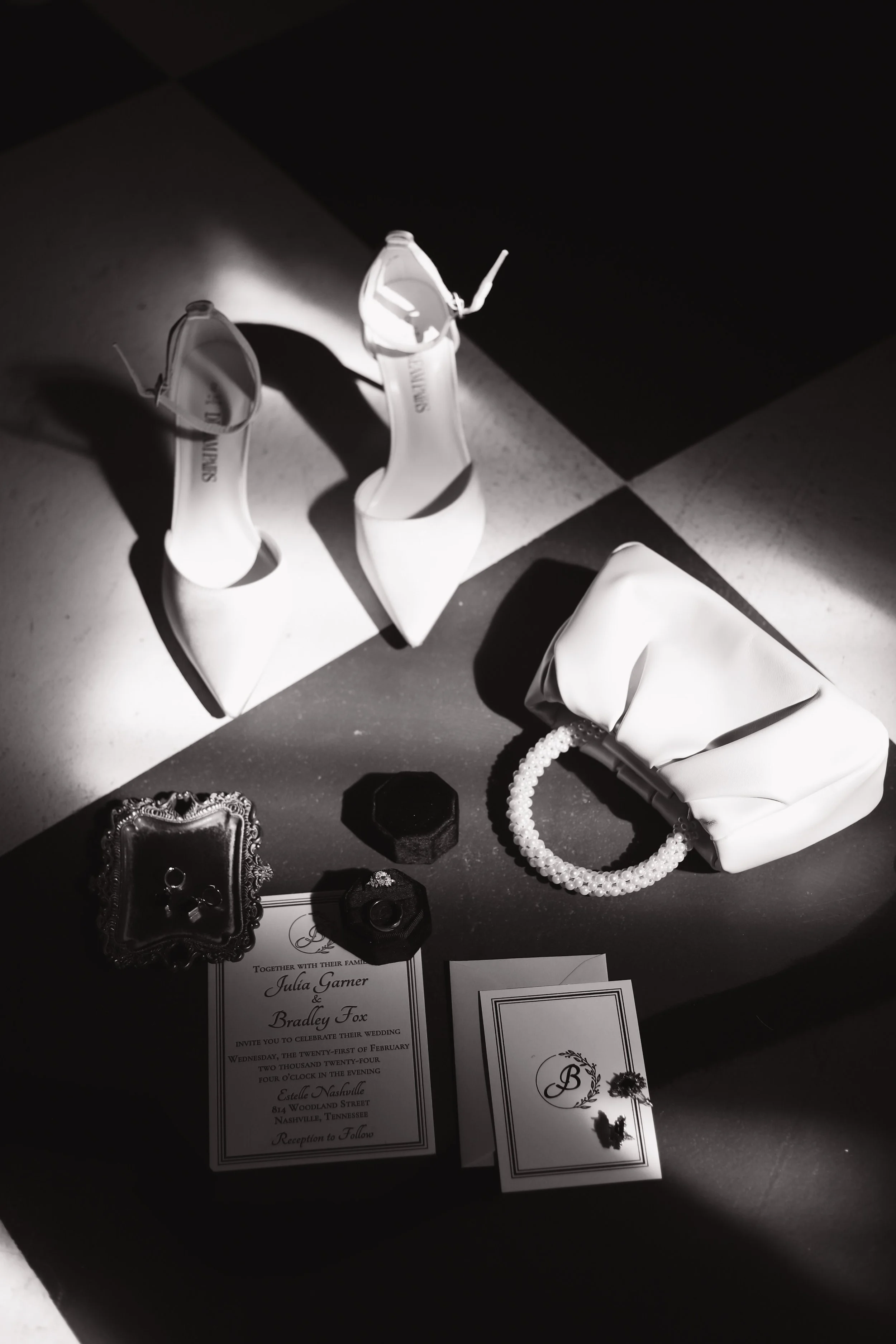 Black and white photo of wedding accessories including high heels, a purse with a pearl handle, an invitation, jewelry, and a ring in a box on a checkered floor.