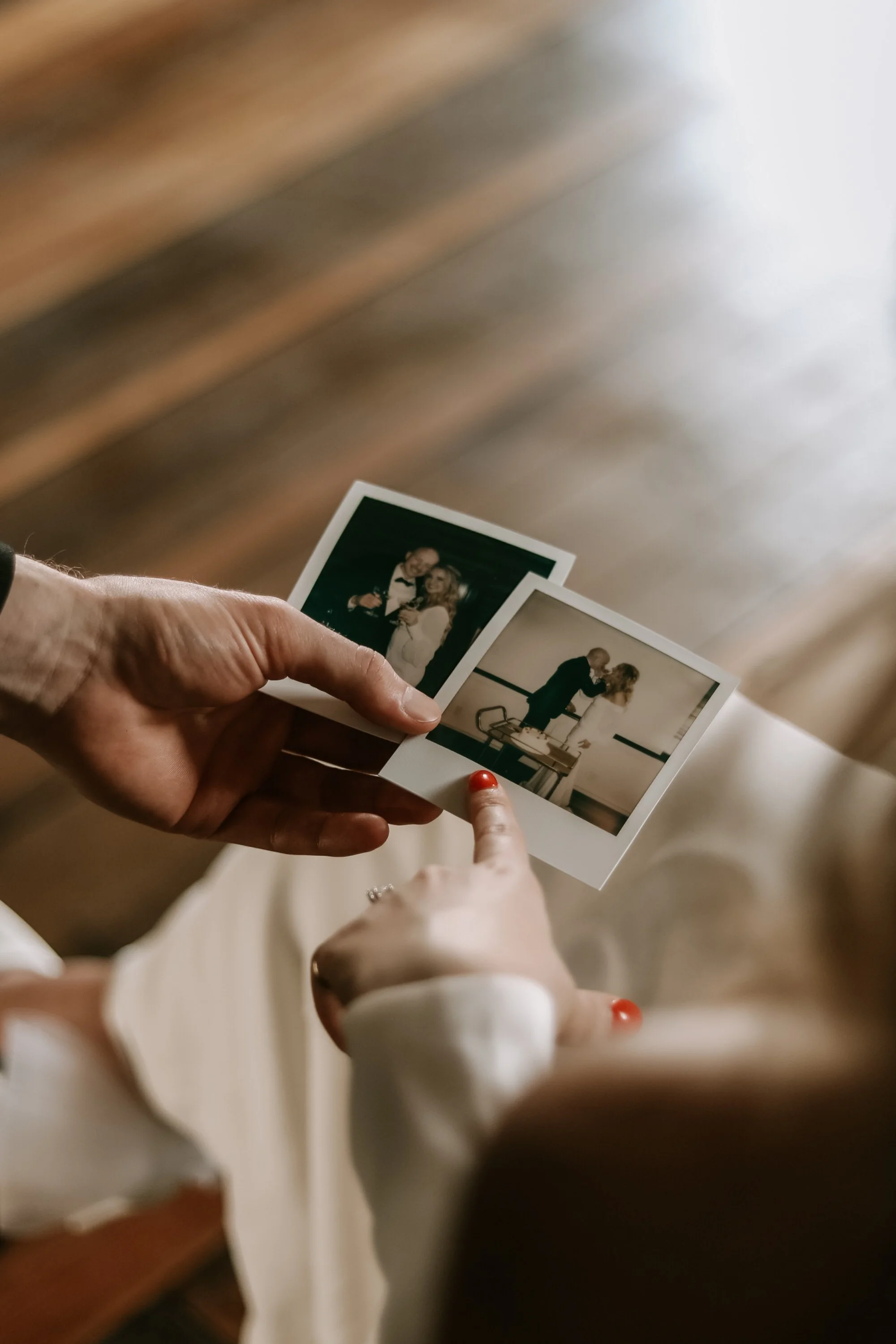 Hands holding two instant photos of a couple celebrating, one kissing at a table, the other smiling with drinks.