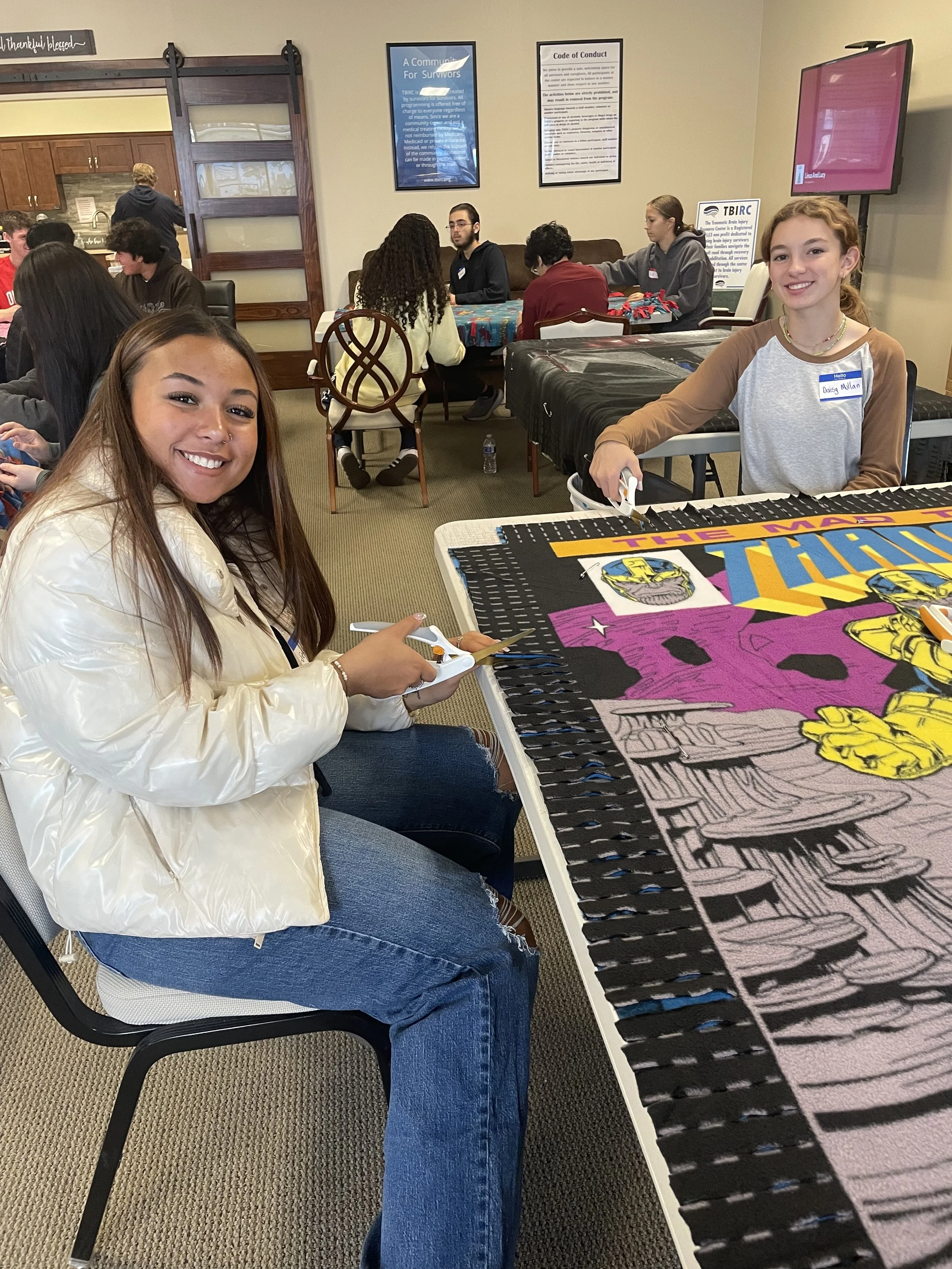 Two YIPEE students sit at a table while cutting fabric to make blankets