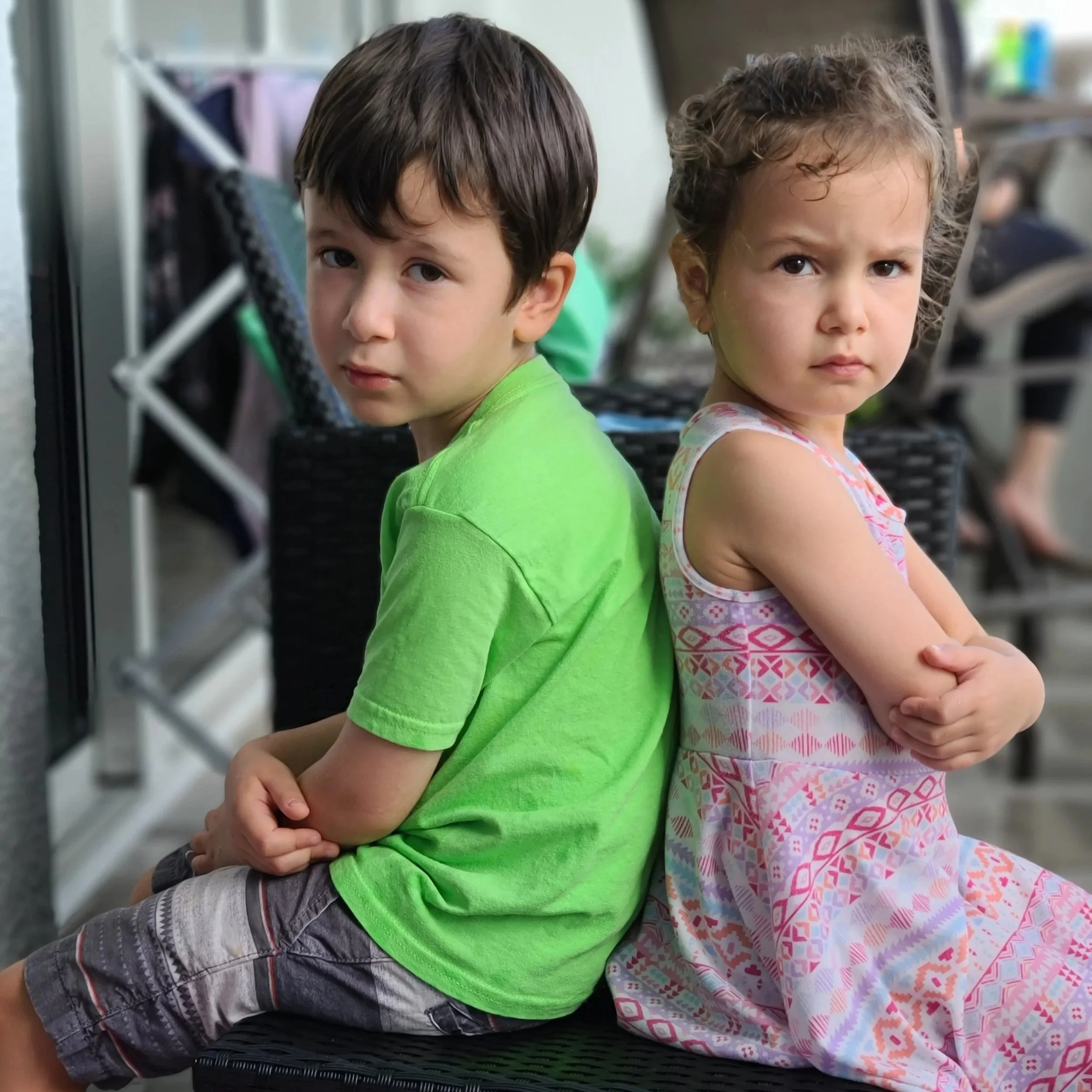 Two young children, a boy and a girl, sit back-to-back on a black woven bench, looking serious and slightly upset. The boy is wearing a bright green shirt and gray shorts and the girl is wearing a sleeveless dress with a pink, orange, and purple pattern.