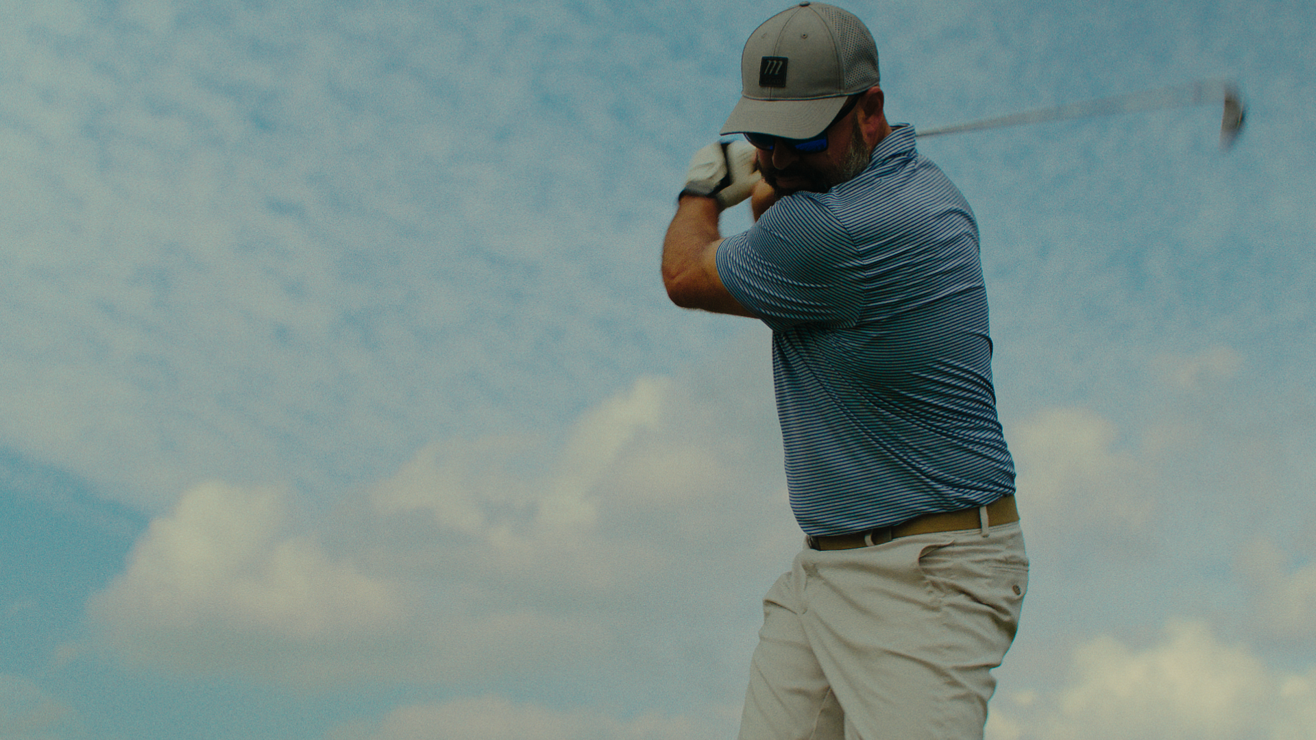 Golfer in a cap and striped polo shirt swinging a golf club against a cloudy sky