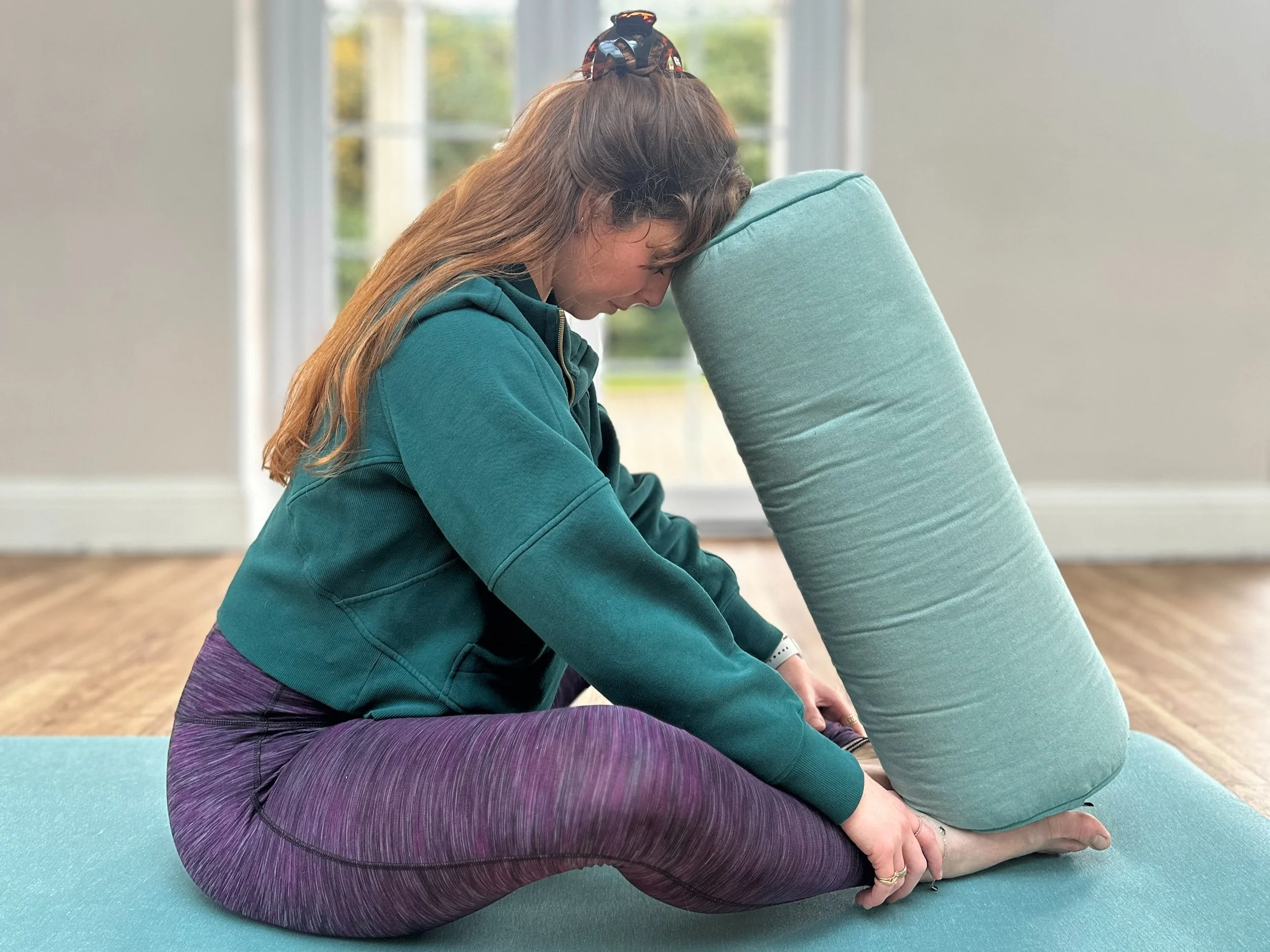 A person practicing yoga indoors, sitting on a yoga mat with legs bent and hugging a cushion for support, wearing a green hoodie and purple leggings.