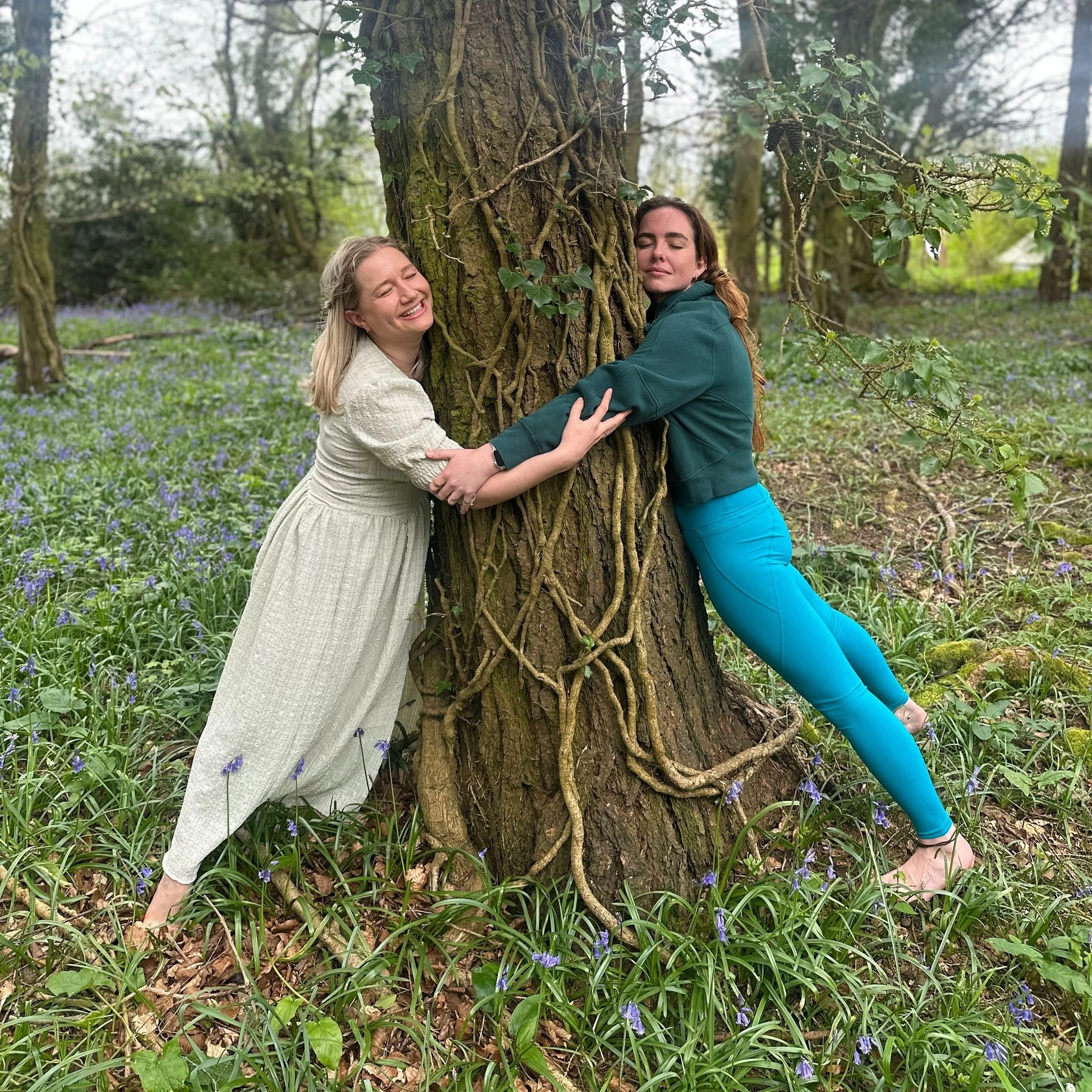 Two women hugging a large tree in a forest surrounded by greenery and purple flowers, smiling and appearing relaxed.